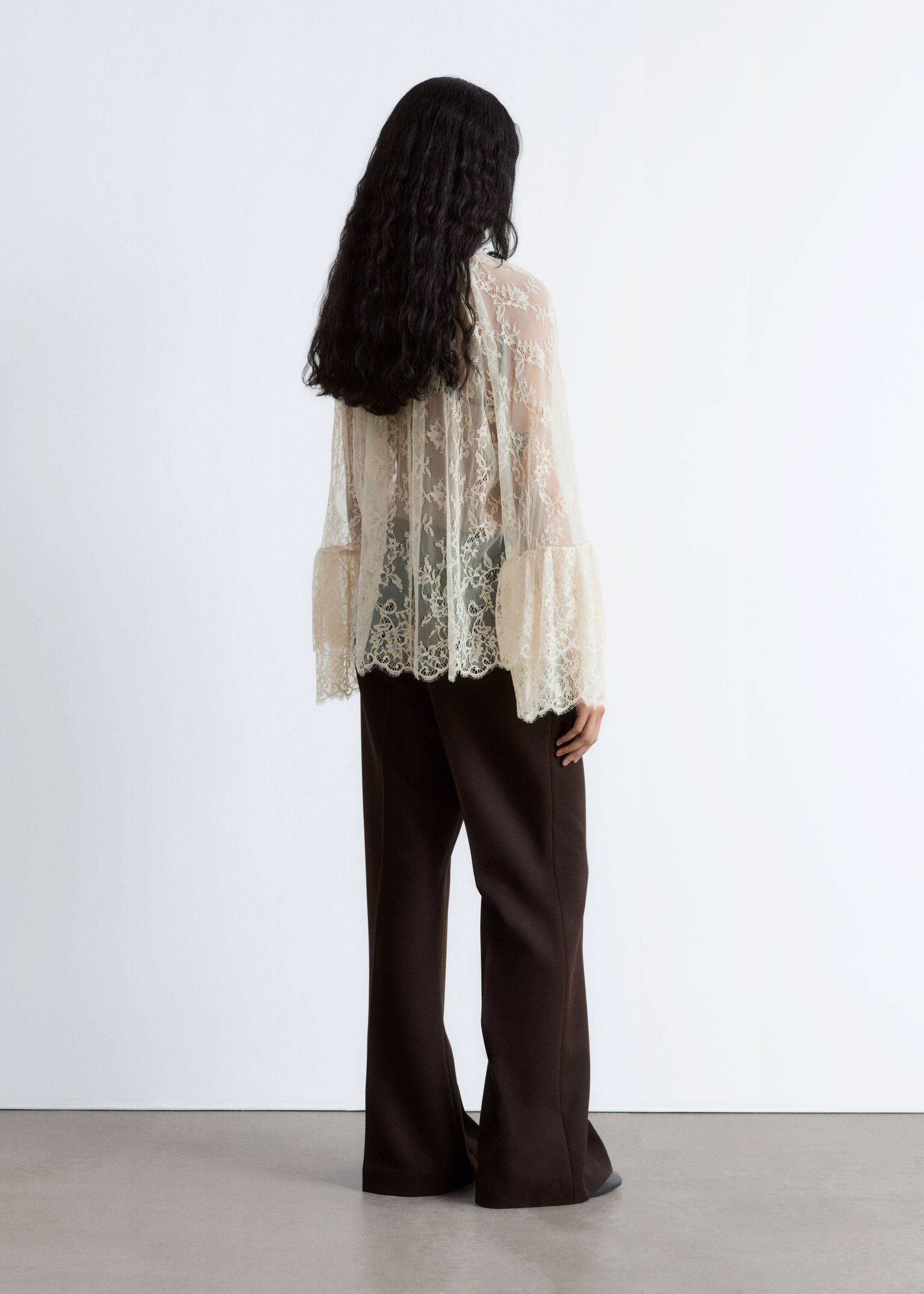 Agrandir l'image: Une blouse en dentelle transparente avec une silhouette aérienne et des drapés autour du cou est portée par le mannequin, avec des manches volumineuses dotées de poignets à double volant et de bords festonnés. La blouse est superposée à un vêtement sombre et associée à un pantalon foncé.