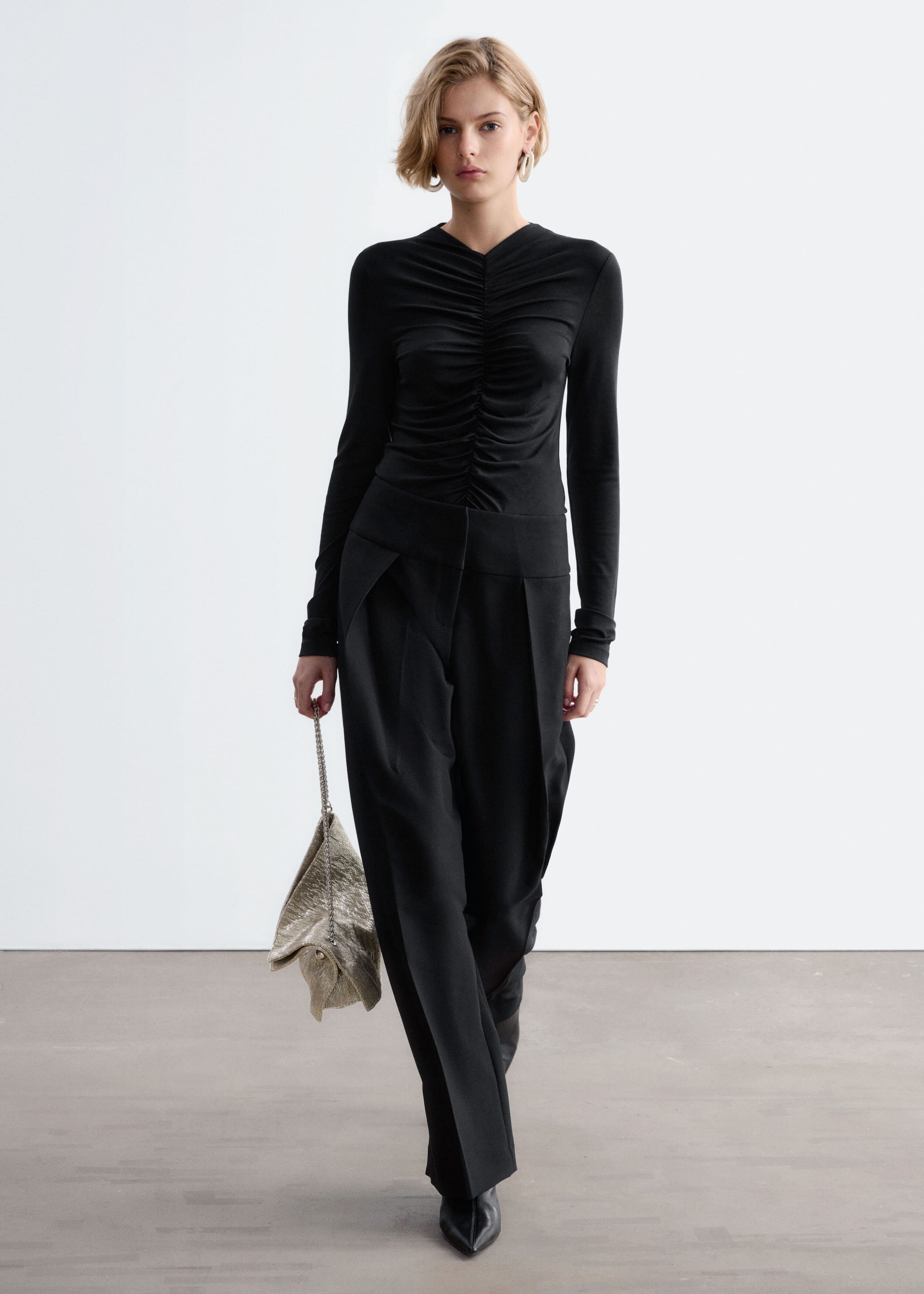 A black, close-fitting, long-sleeved top with ruched gathering along the center front seam is worn by the model. The top is tucked into high-waisted black trousers, and the model is also carrying a metallic gold handbag.