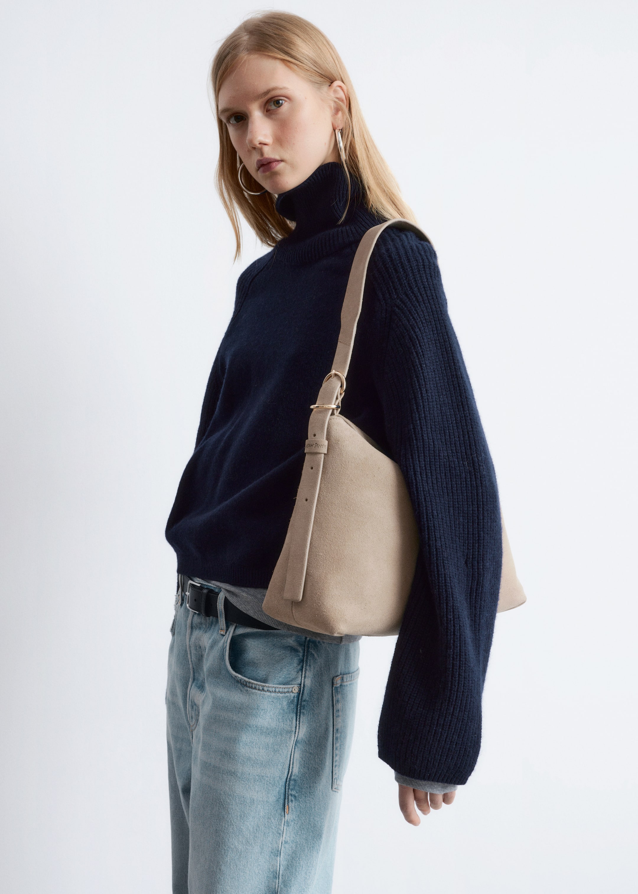 Se større billede: En marineblå turtleneck sweater vises fra en front-venstre perspektiv og har en afslappet pasform med brede, ribbede ærmer og en foldet hals. En lys grå t-shirt lagdeles nedenunder, og en beige skuldertaske hviler over sweaterens front.