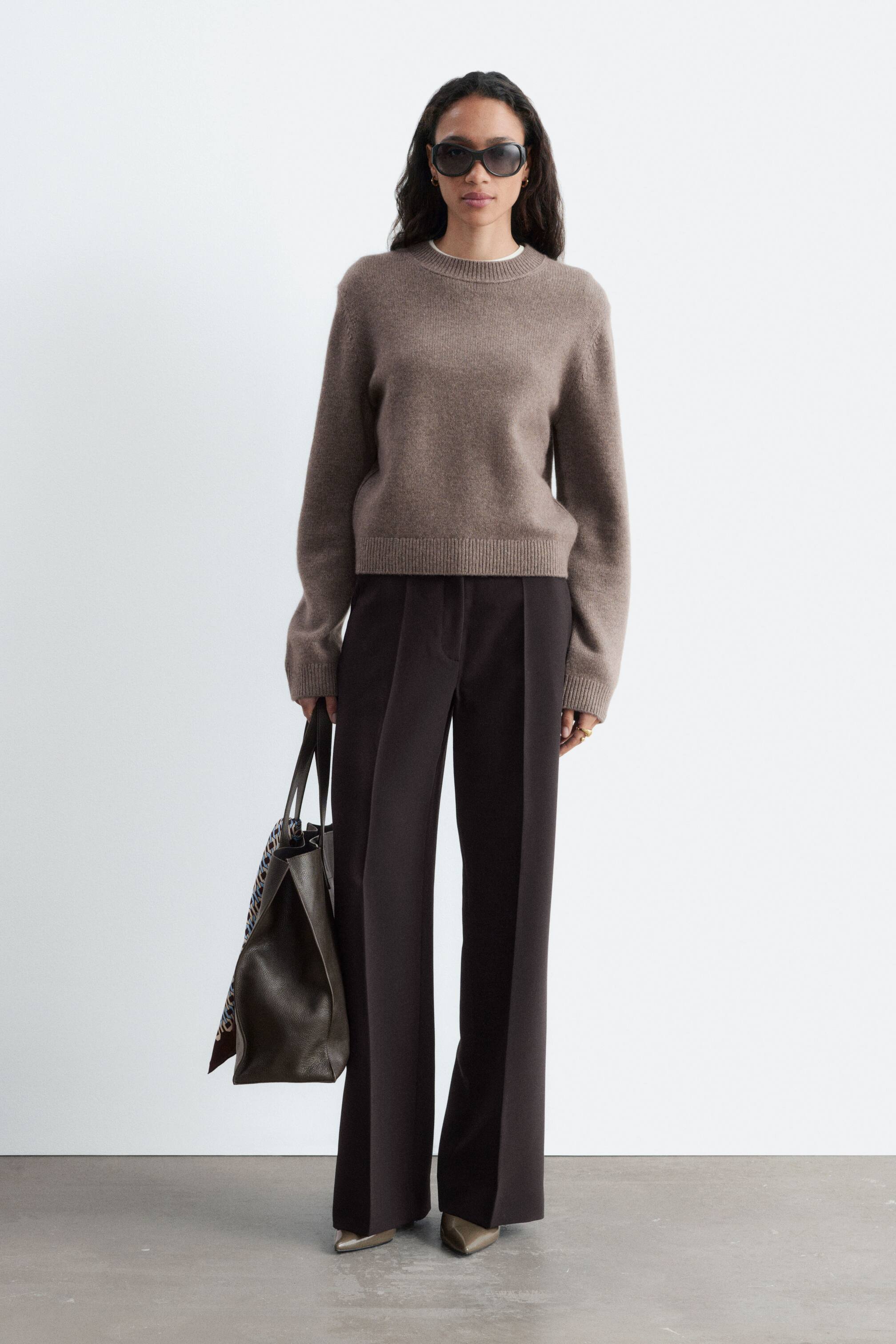 View larger image: Dark brown loose-fit trousers featuring a regular waist and sharp front press creases are worn by the model. The trousers are paired with a taupe crew-neck sweater, pointed-toe heels, and a dark leather tote bag that the model is carrying.