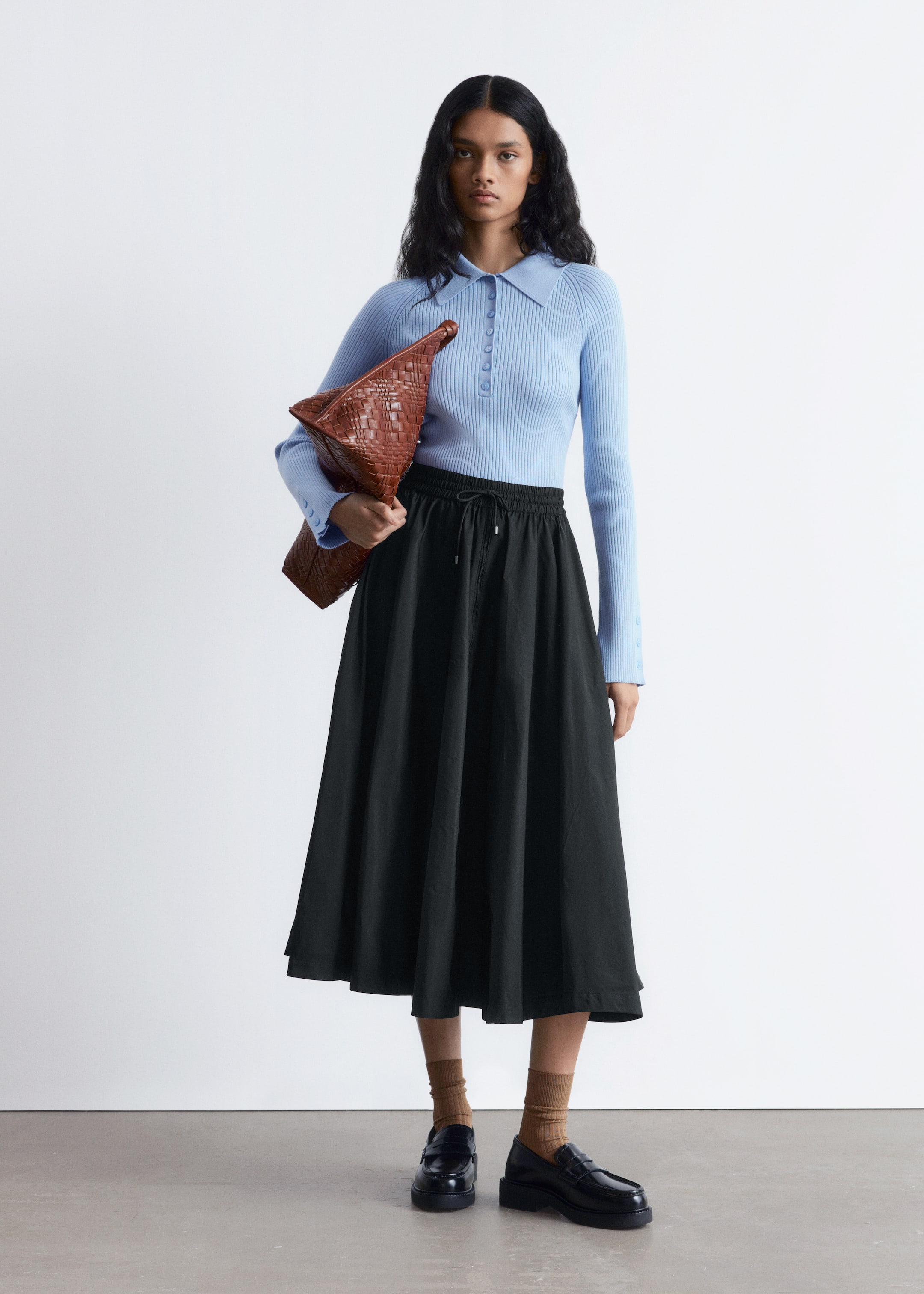 Agrandir l'image: Un haut polo en tricot côtelé bleu clair avec une patte de boutonnage est porté, rentré dans une jupe midi noire ample avec une taille élastiquée à cordon. L'ensemble est complété par des chaussettes côtelées marron à la cheville, des mocassins noirs épais et une pochette tressée marron tenue à la main.