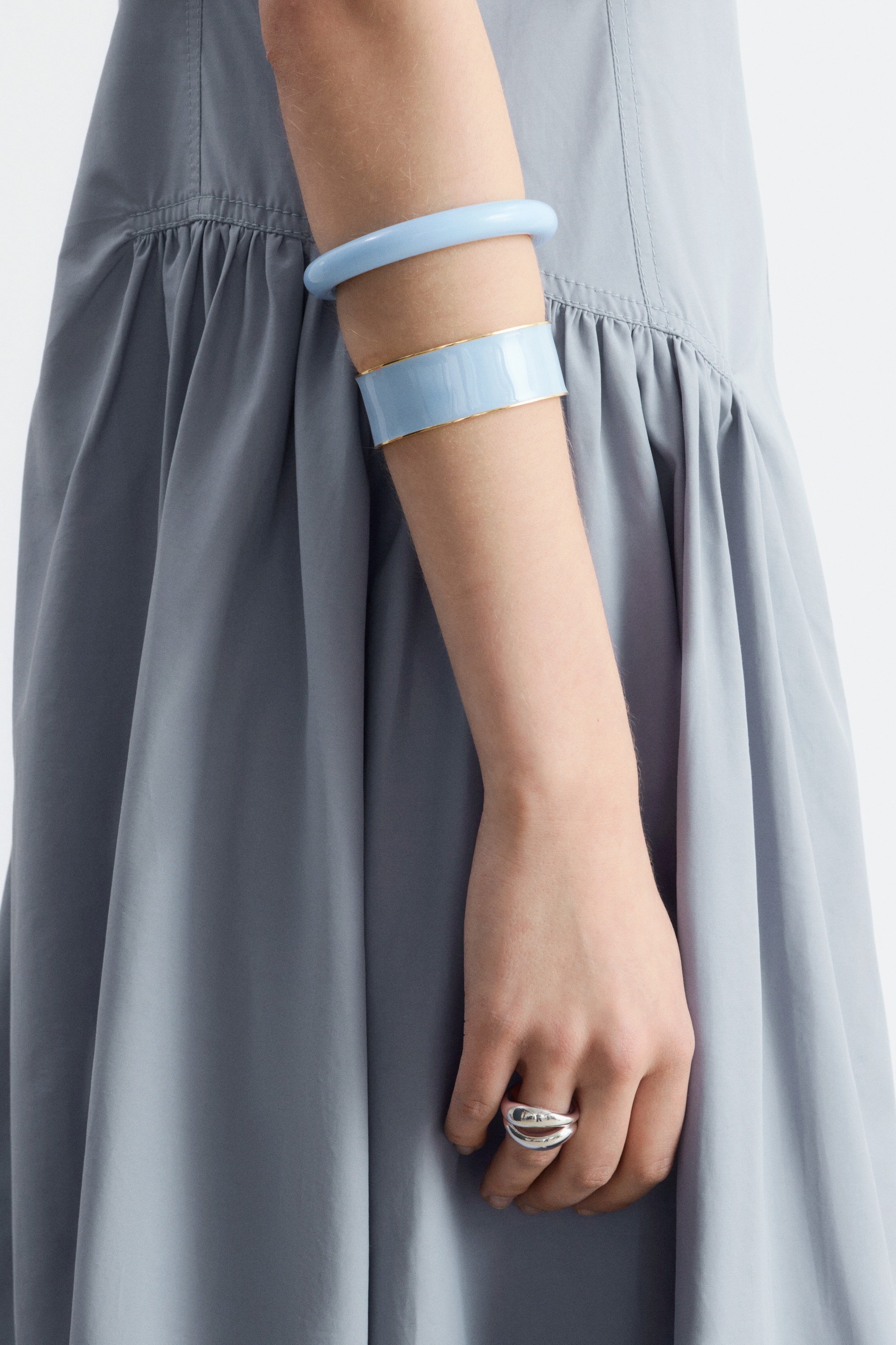 Agrandir l'image: Un bracelet épais et arrondi de couleur bleu clair et un large bracelet manchette émaillé bleu clair avec un bord doré sont portés sur un bras, tandis qu'une bague multi-bandes argentée est portée sur un doigt.
