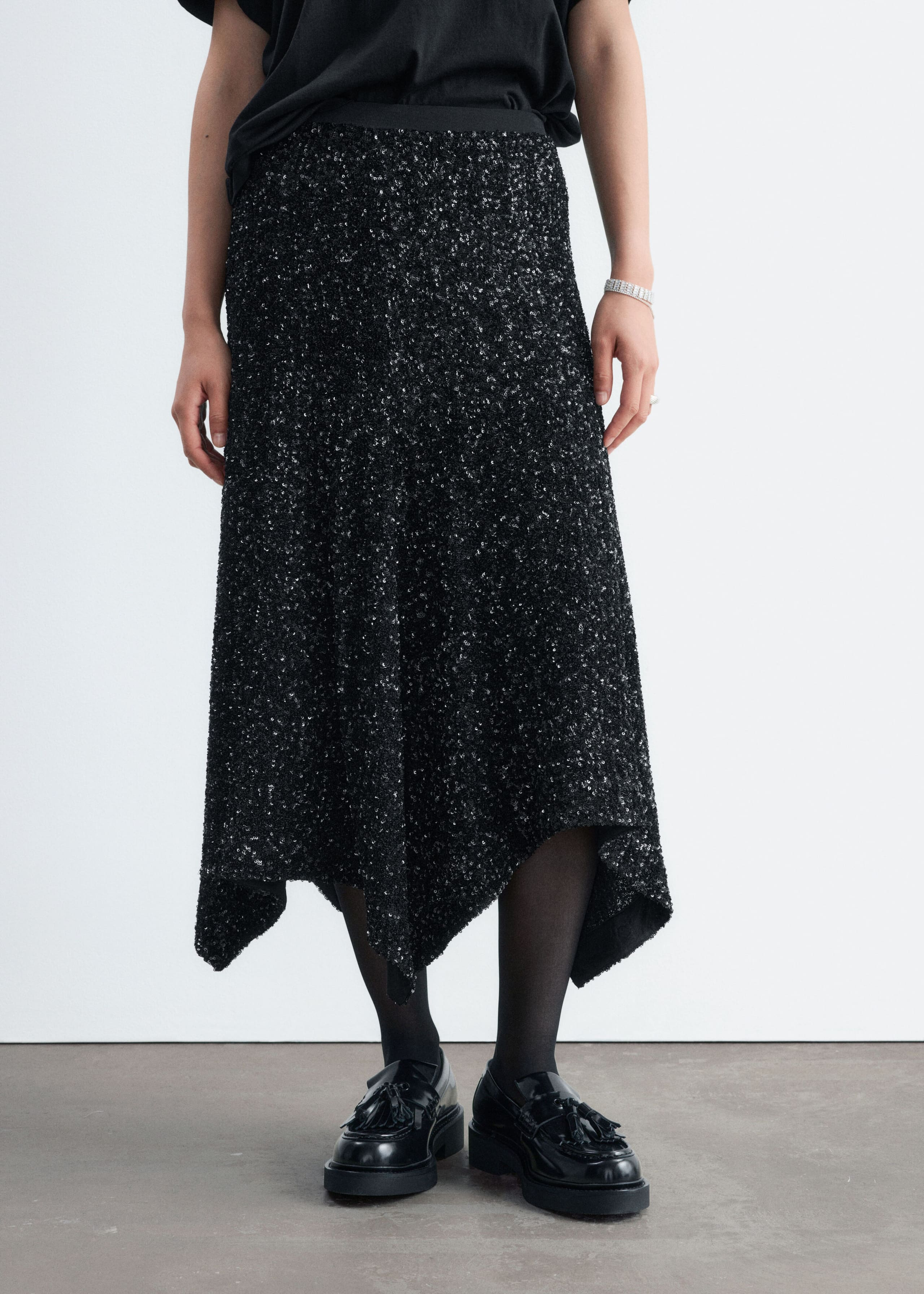 A black sequined skirt with an asymmetric hem is being worn by the model. The skirt is styled with a tucked-in black T-shirt, sheer black tights, and black patent leather loafers.