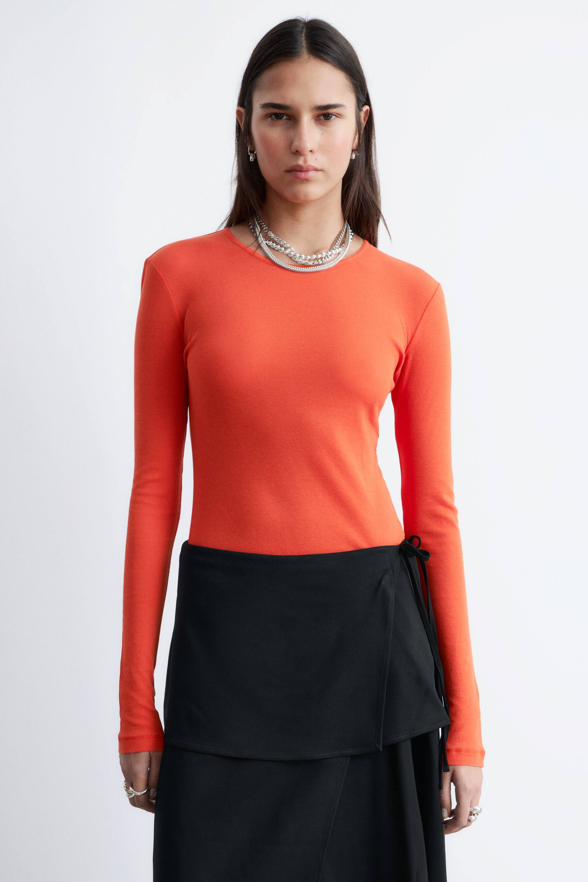 View larger image: A vibrant orange, fitted long-sleeved top is displayed from the front, showcasing a smooth texture. Its crew neck is partially adorned with layered necklaces, and the top is worn tucked in at the waist, accentuating its slim fit.