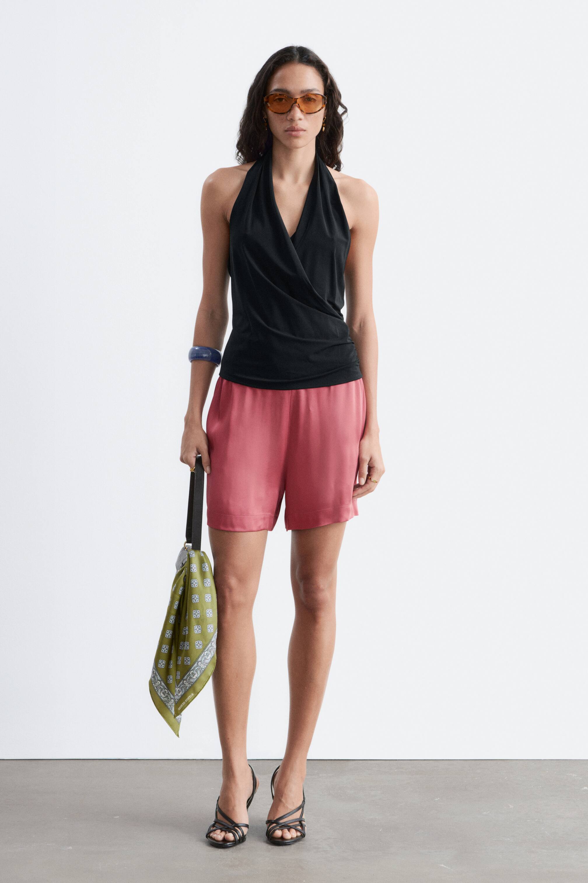 Se større billede: Rosa satinshorts udviser en glansfuld finish, en afslappet silhuet og en synlig elastisk høj talje. Modellen bærer disse shorts og styler dem med en sort halterneck-top og sorte sandaler med remme.