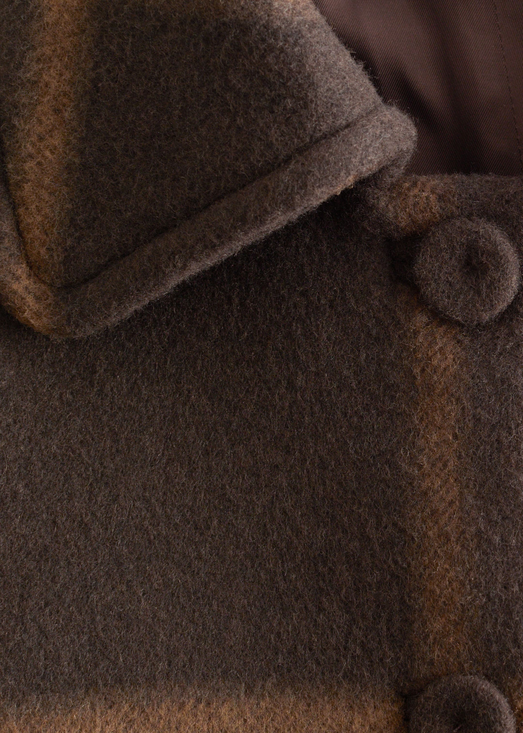 Grotere afbeelding bekijken: Een wazig wollen kledingstuk toont zijn donkerbruine en beige gestreepte textuur. Het heeft een dikke, opgerolde rand aan de linkerbovenkant, een bijpassende met stof bedekte knoop aan de rechterkant en een gladde, donkerbruine voering die zichtbaar is onder de opgerolde rand.