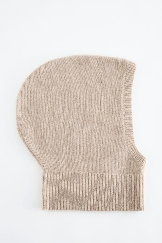 A beige knitted balaclava hood is lying flat in side profile, showcasing its fine-knit texture, a ribbed trim framing the face opening, and a wide ribbed band at the neck. - 1