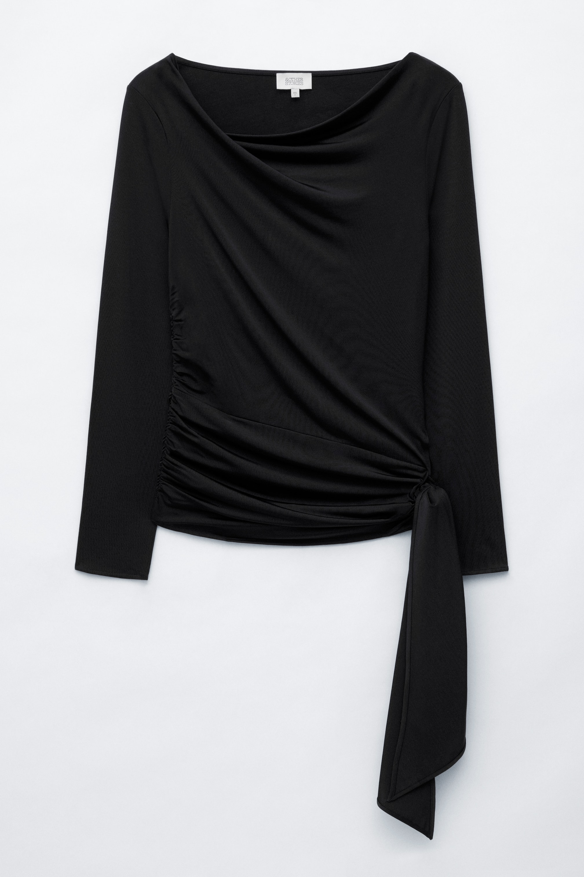 Vezi imaginea mai mare: Un top negru din jerseu cu mâneci lungi este prezentat din față, având un decolteu asimetric, drapat și drapaj pe o parte. Designul este completat de un detaliu lung, din material textil, care atârnă de la talia fronsată.