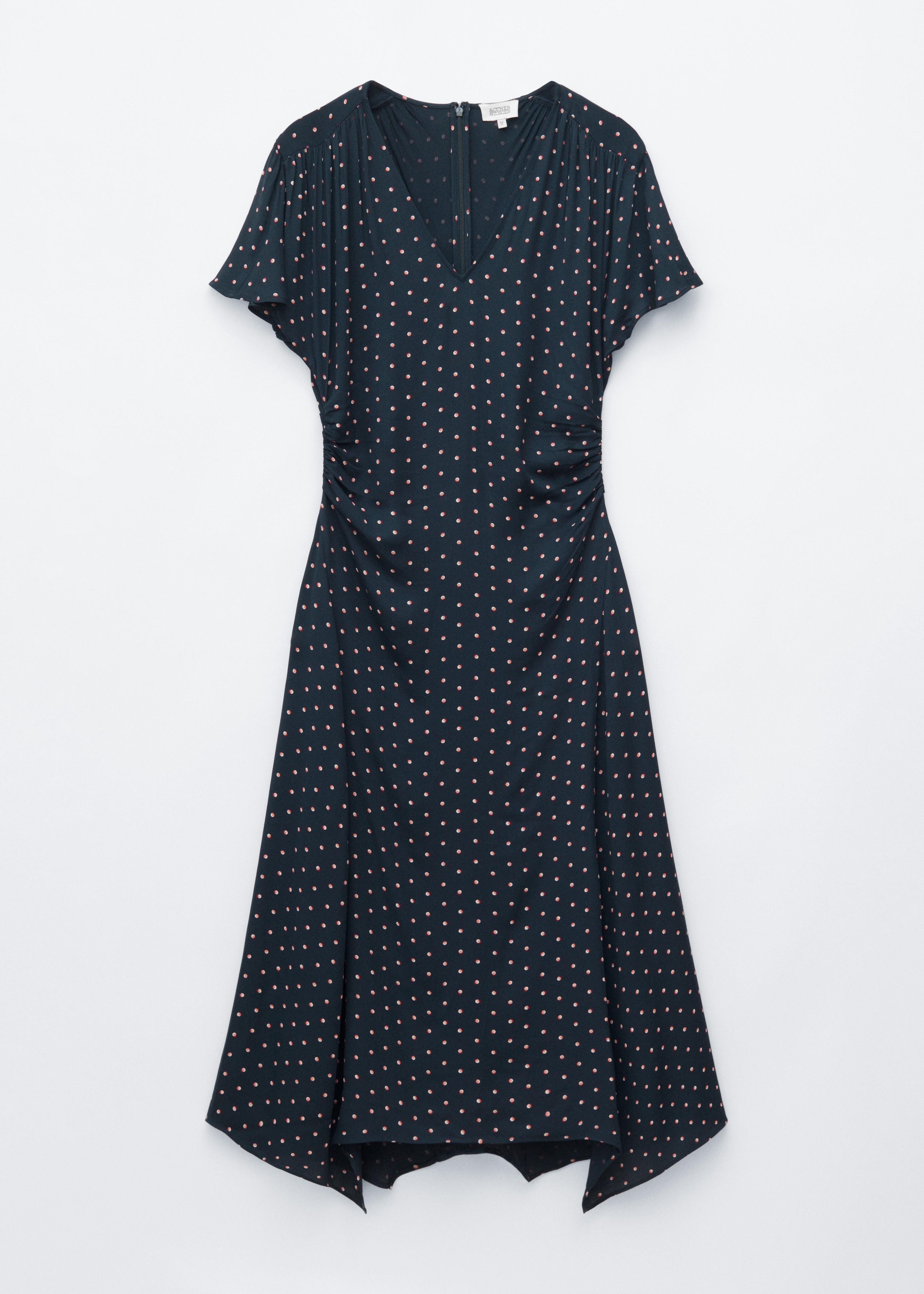 Une robe midi bleu foncé à pois roses est portée par le mannequin, avec un col en V, des manches courtes et de légers fronces aux épaules et à la taille. Un pull marron est noué à la taille de la robe, et le mannequin porte un sac à main marron foncé.