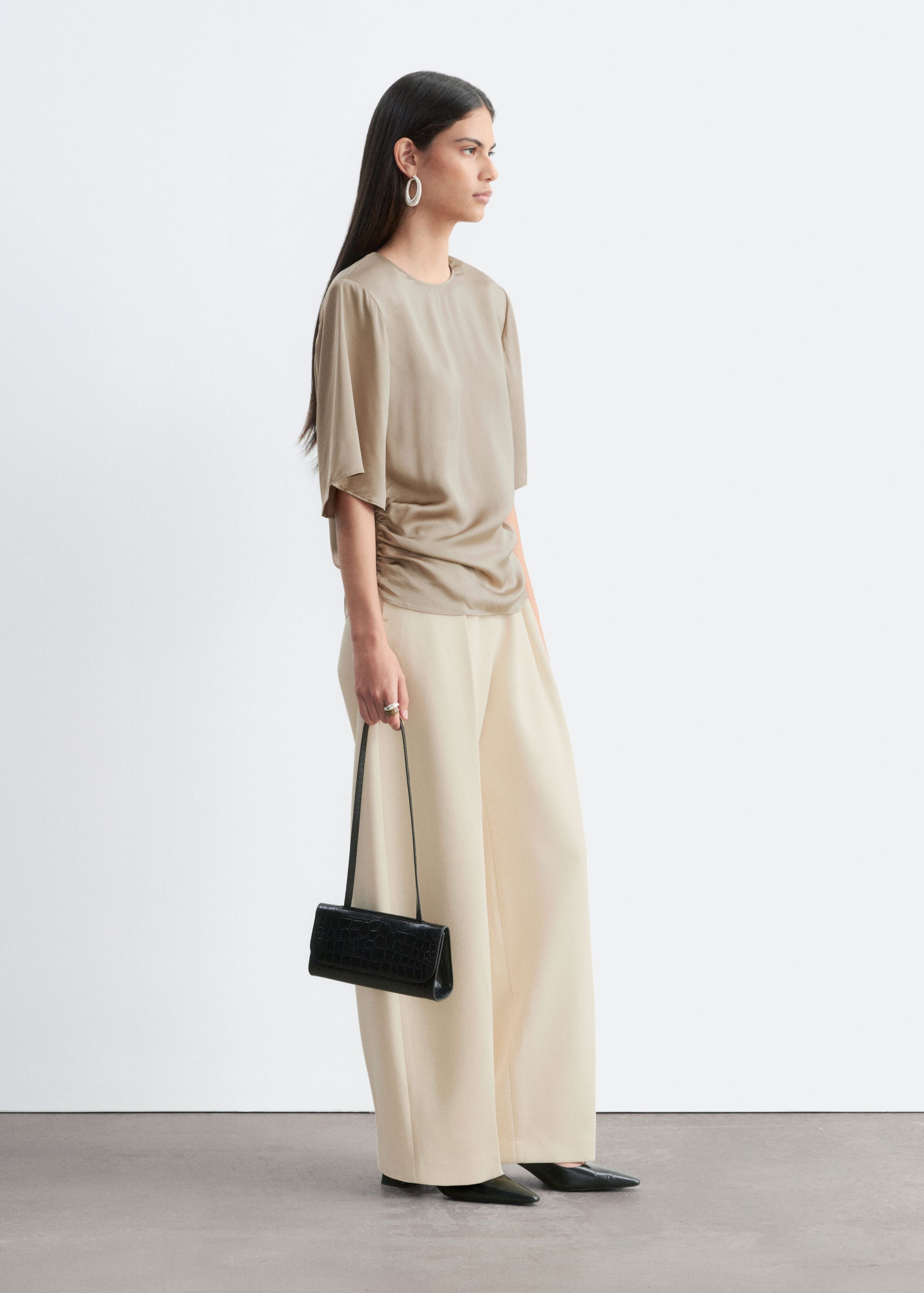 A beige short-sleeve blouse featuring soft shoulder pads and gathered side pleats is being worn by the model. The blouse is styled with cream-colored wide-leg trousers, and the model is holding a small black shoulder bag.