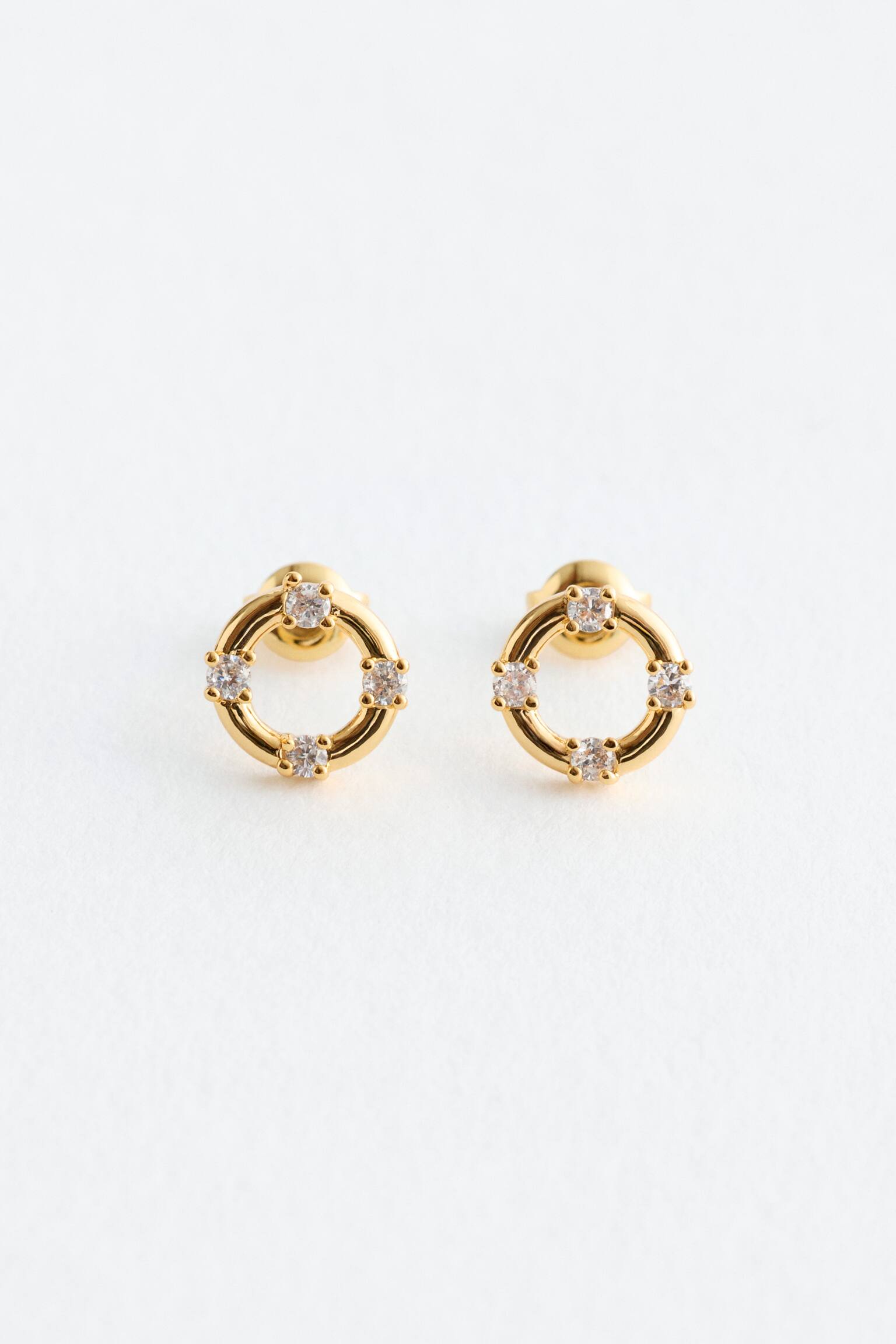 Une paire de boucles d'oreilles à clous de couleur dorée est orientée vers l'avant, chacune présentant un design circulaire avec quatre inserts en strass transparents. - 1