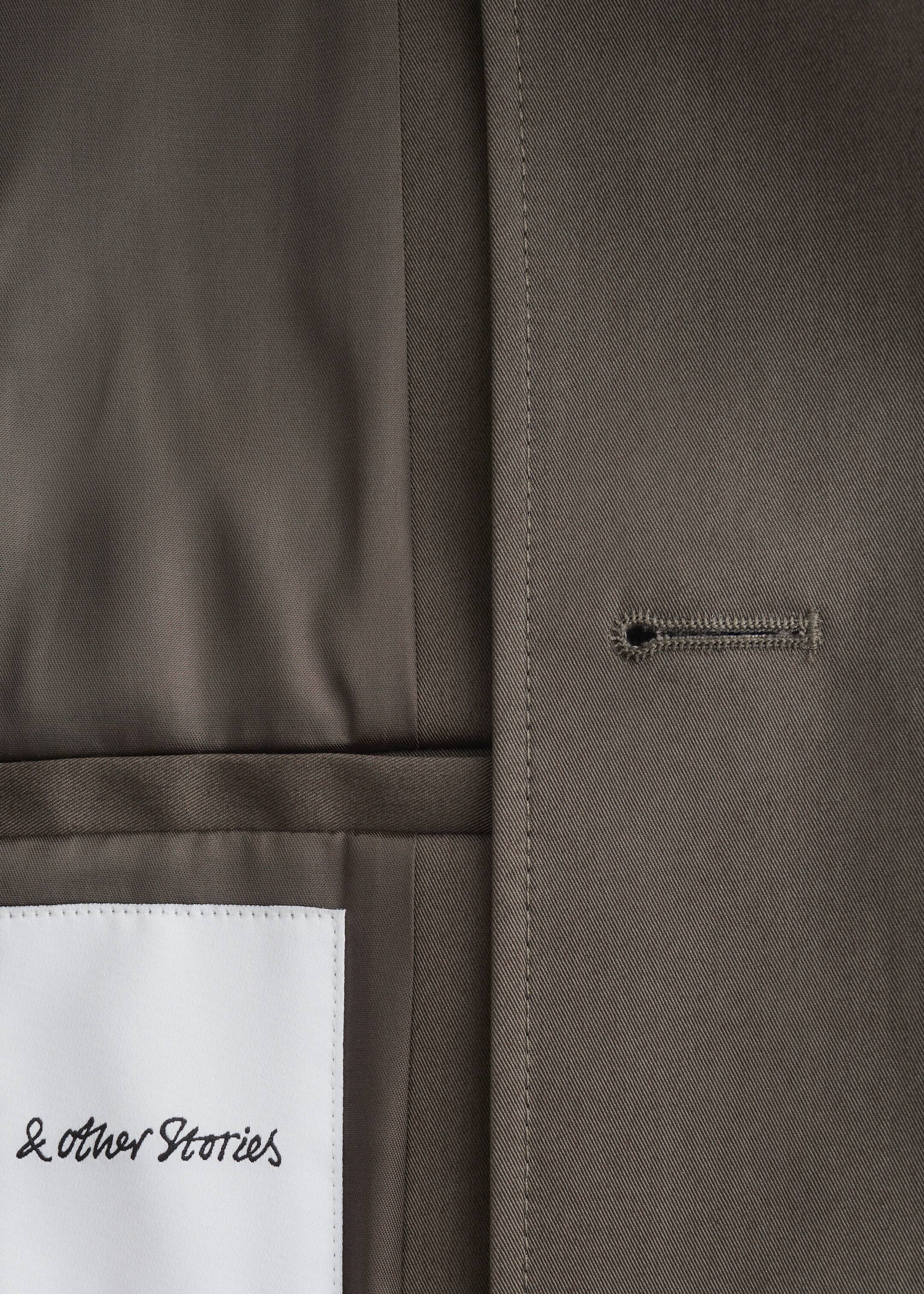 Agrandir l'image: Un gros plan de l'avant d'une veste montre son tissu sergé structuré olive-gris avec un tissage diagonal distinct. Une boutonnière horizontale avec des coutures visibles se trouve à droite, à côté d'une couture verticale avec un surpiquage prononcé. Une étiquette de marque blanche '& Other Stories' est cousue sur le tissu en bas à gauche, révélant une doublure intérieure plus lisse.