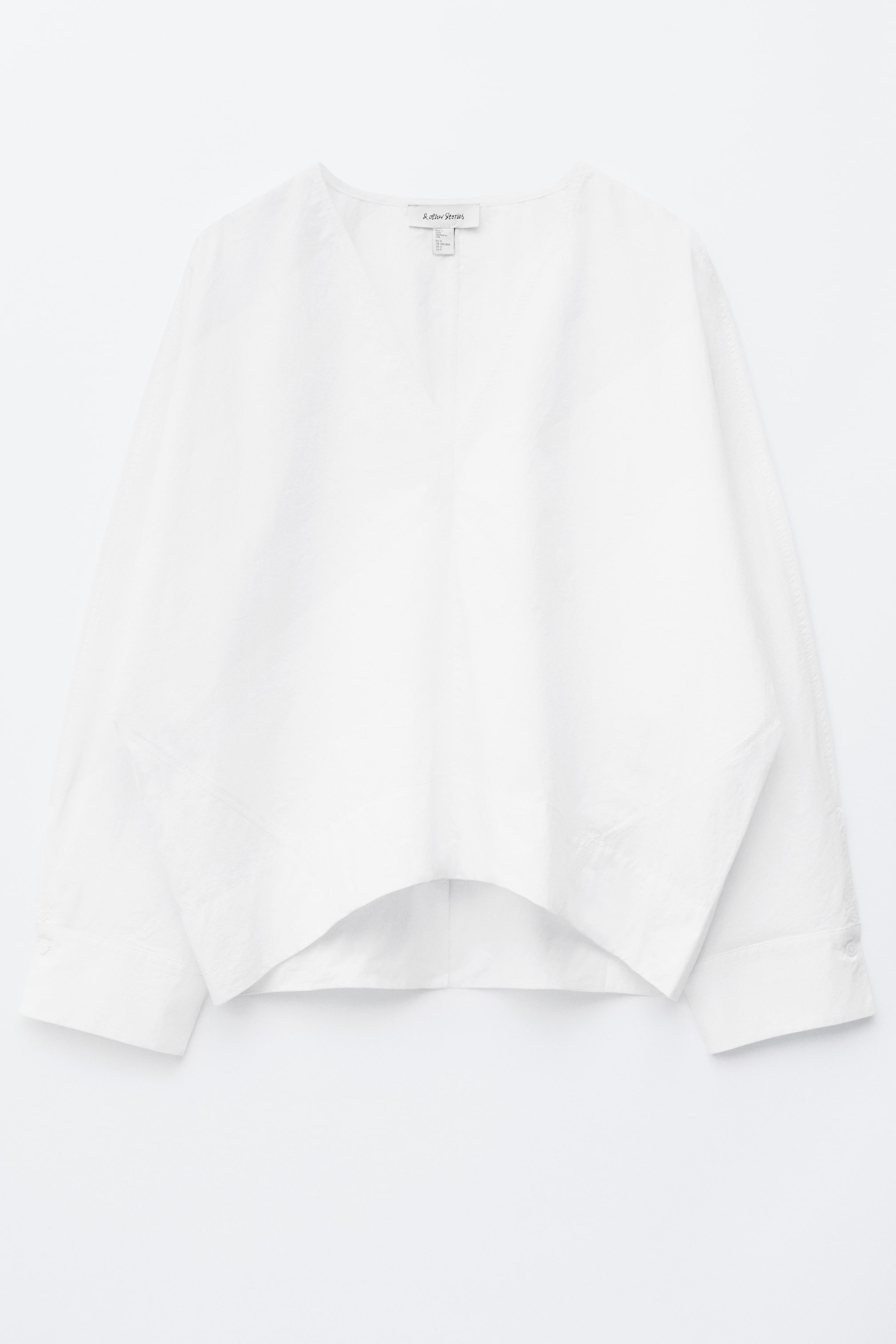 Visa större bild: En vit bomullsblus presenteras framifrån, med en V-ringning och långa, voluminösa kokongformade ärmar med knäppta manschetter. Den visar upp en skulptural silhuett med en böjd fåll.