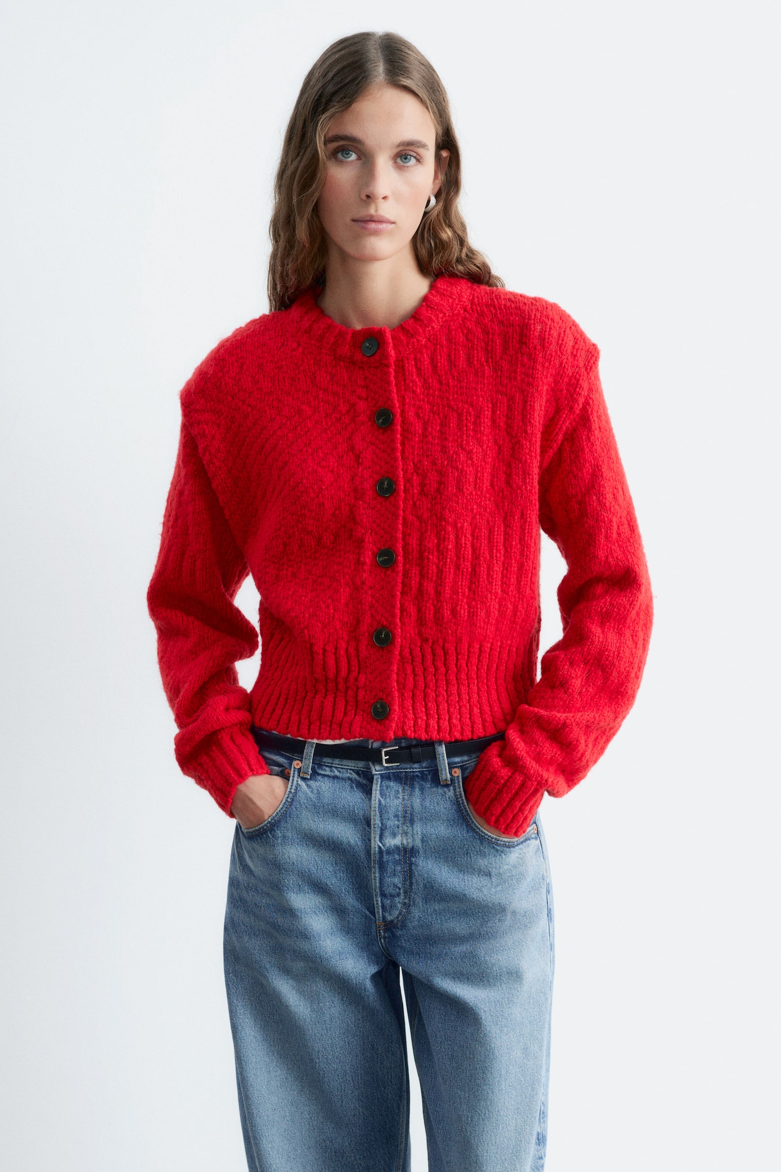 En rød cardigan i ull og bomull med strukturstrikk, knappelukking foran med mørke knapper og rund hals med ribbestrikk bæres av modellen. Cardiganen styles med blå jeans, og modellen har hendene hvilende i de fremre lommene på buksene. - 1