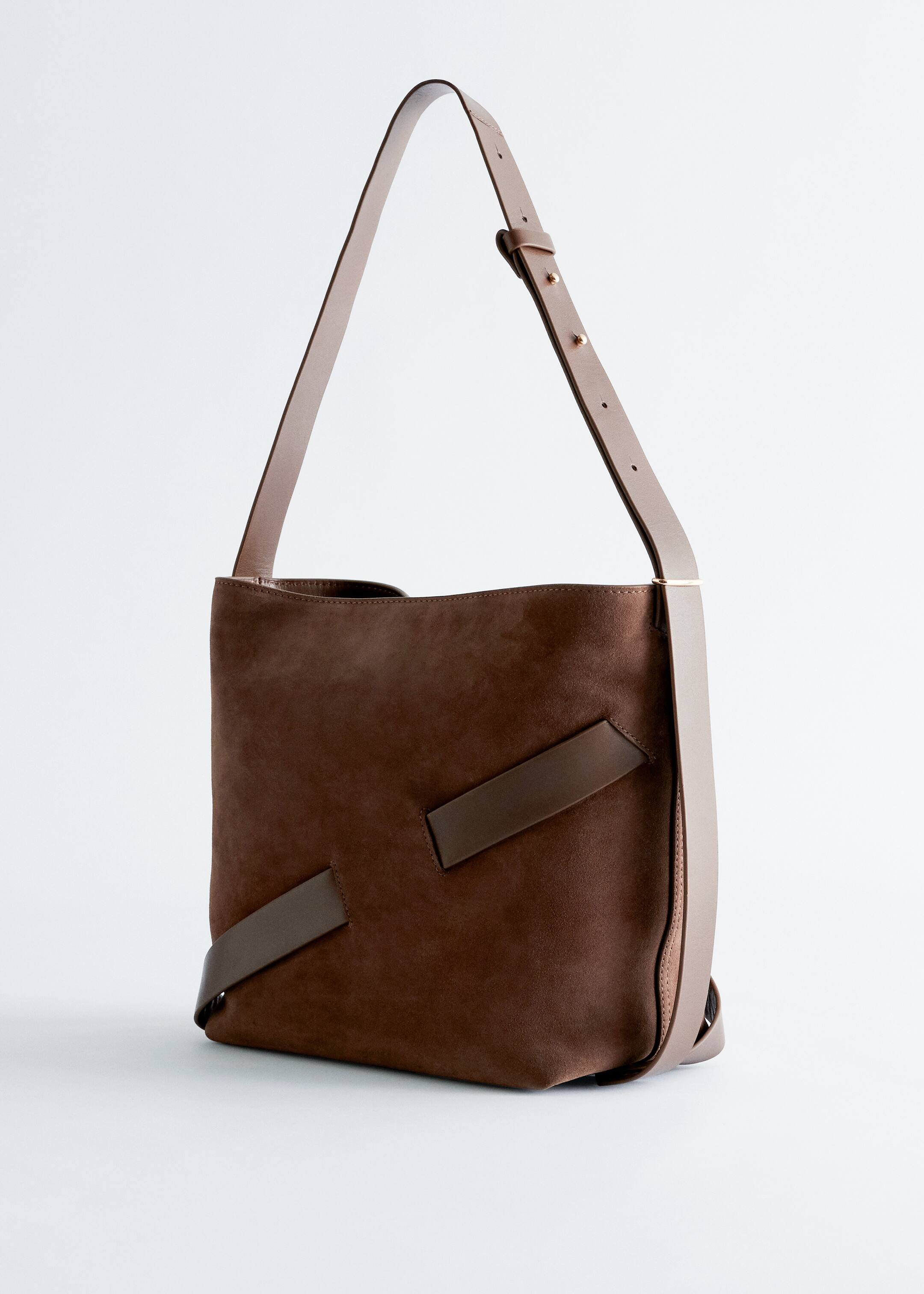 Grösseres Bild ansehen: Eine braune Bucket Bag präsentiert sich in einem leichten linken Winkel und hat einen Wildlederkörper mit dunkelbraunen Ledergriffen, die diagonal auf der Vorderseite platziert sind. Hellere, hellbraune Lederbesätze sind an den Seiten sichtbar, und ein verstellbarer, hellbrauner Lederschulterriemen mit goldfarbenen Nieten erstreckt sich von der Tasche nach oben.