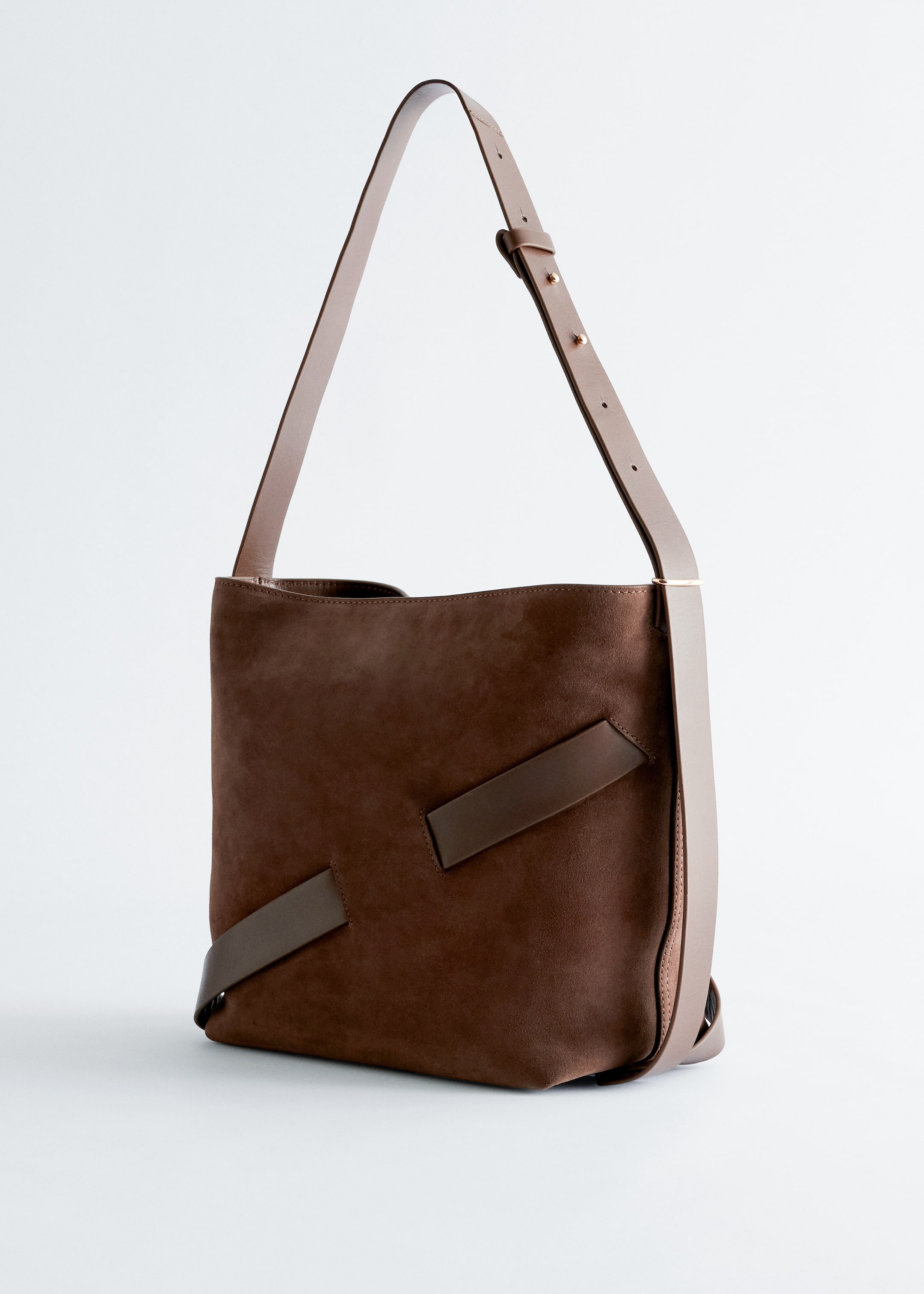 View larger image: A brown bucket bag is presenting at a slight left angle, featuring a suede body with darker brown leather straps diagonally placed on its front. Lighter tan leather trims are visible on its sides, and an adjustable lighter tan leather shoulder strap with gold-tone studs is extending upwards from the bag.