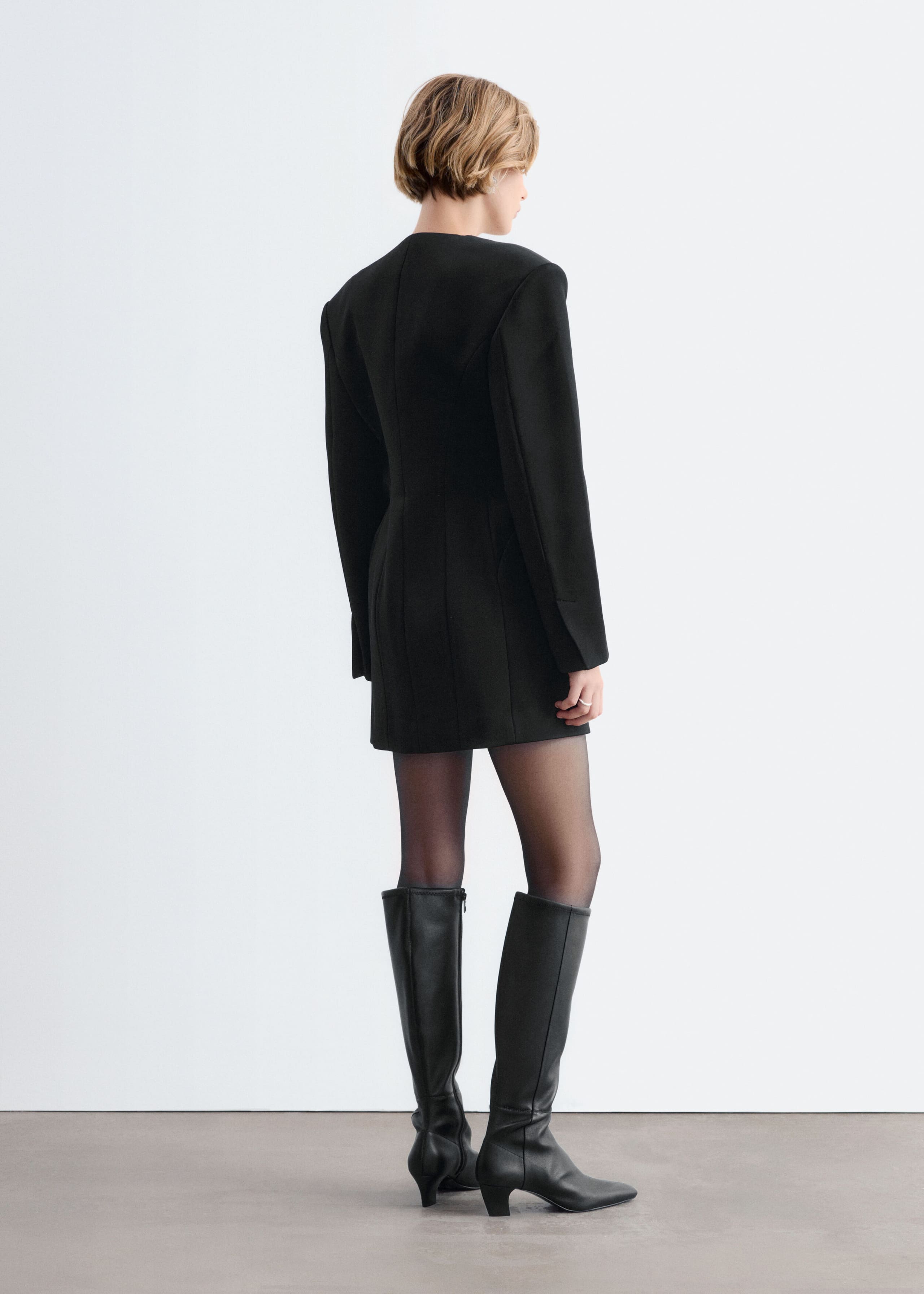 A black blazer dress with structured shoulders, a cinched waist, and vertical seams is worn by the model in a back view. The long-sleeved dress is styled with sheer black tights and black leather knee-high boots.