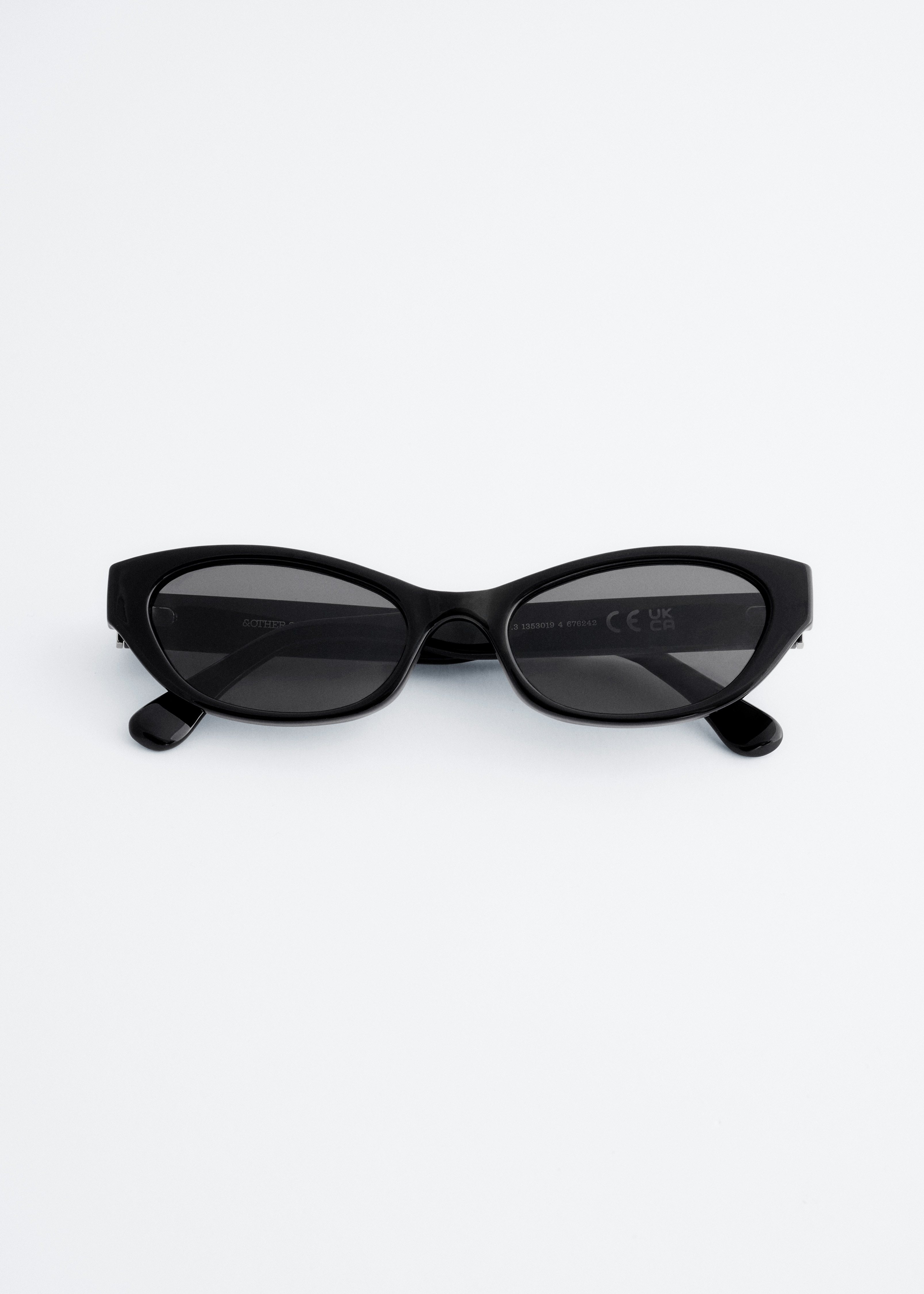 Uns Ã³culos de sol estreitos e ovais, com armaÃ§Ãµes escuras e lentes fumadas, estÃ£o a ser usados pelo modelo. Os Ã³culos de sol sÃ£o combinados com um casaco texturado castanho-escuro.