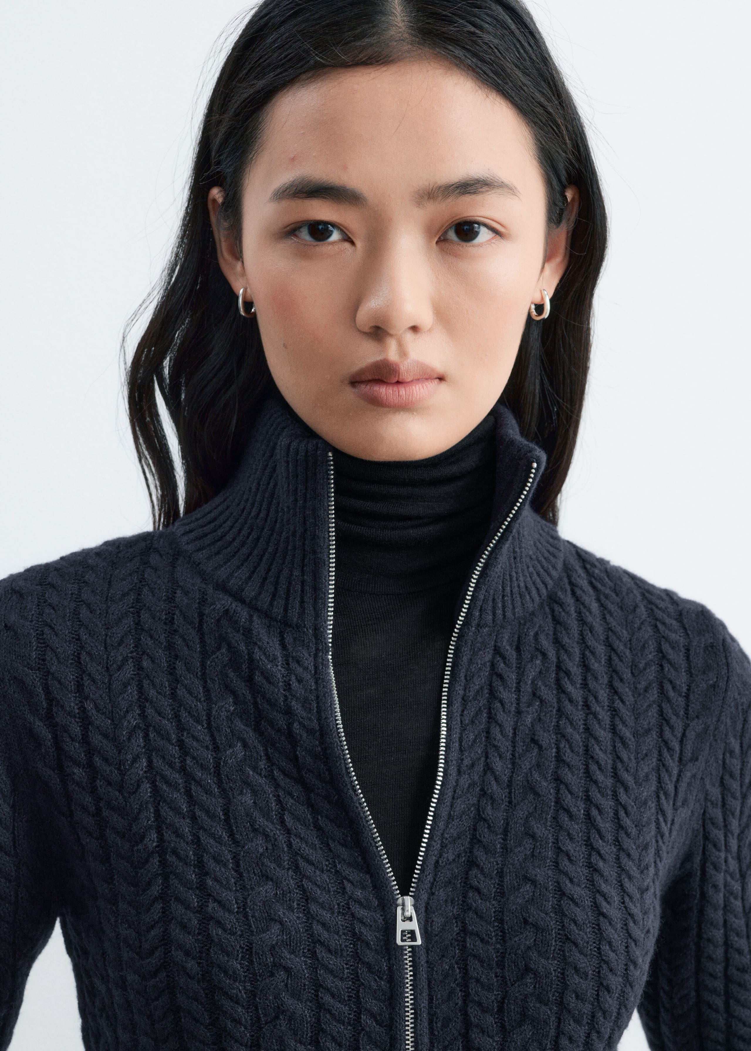 A dark grey cable-knit cardigan with a high ribbed collar and a silver front zip is being worn by the model. The cardigan is partially unzipped, showing a black roll-neck top layered underneath.