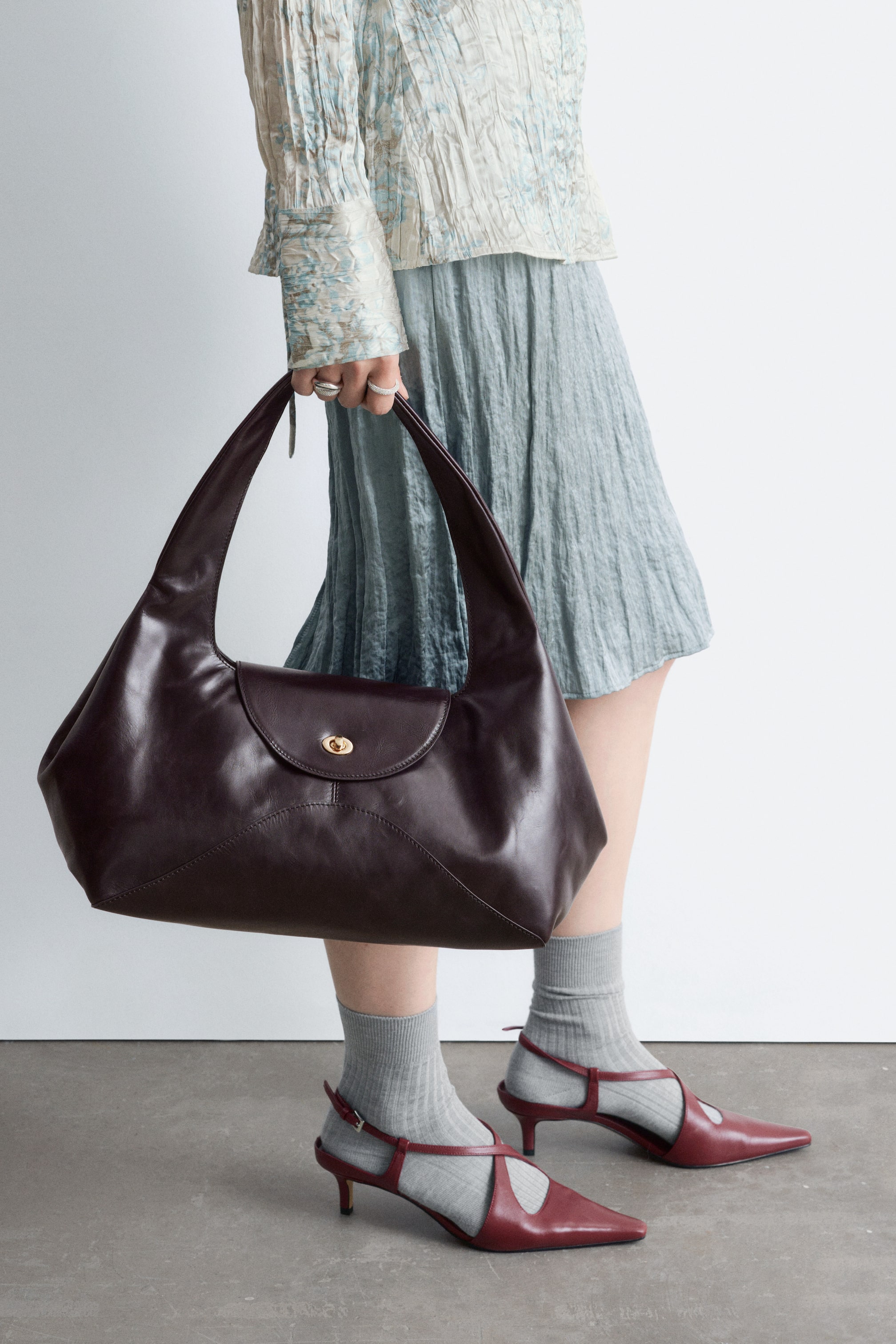 Visa större bild: En mörkbrun axelväska i läder, med en böjd, skulptural silhuett, hålls av modellen. Väskan har ett brett, sluttande axelband, en frontklaff med vridlåsförslutning och en dekorativ skärlinjesöm.