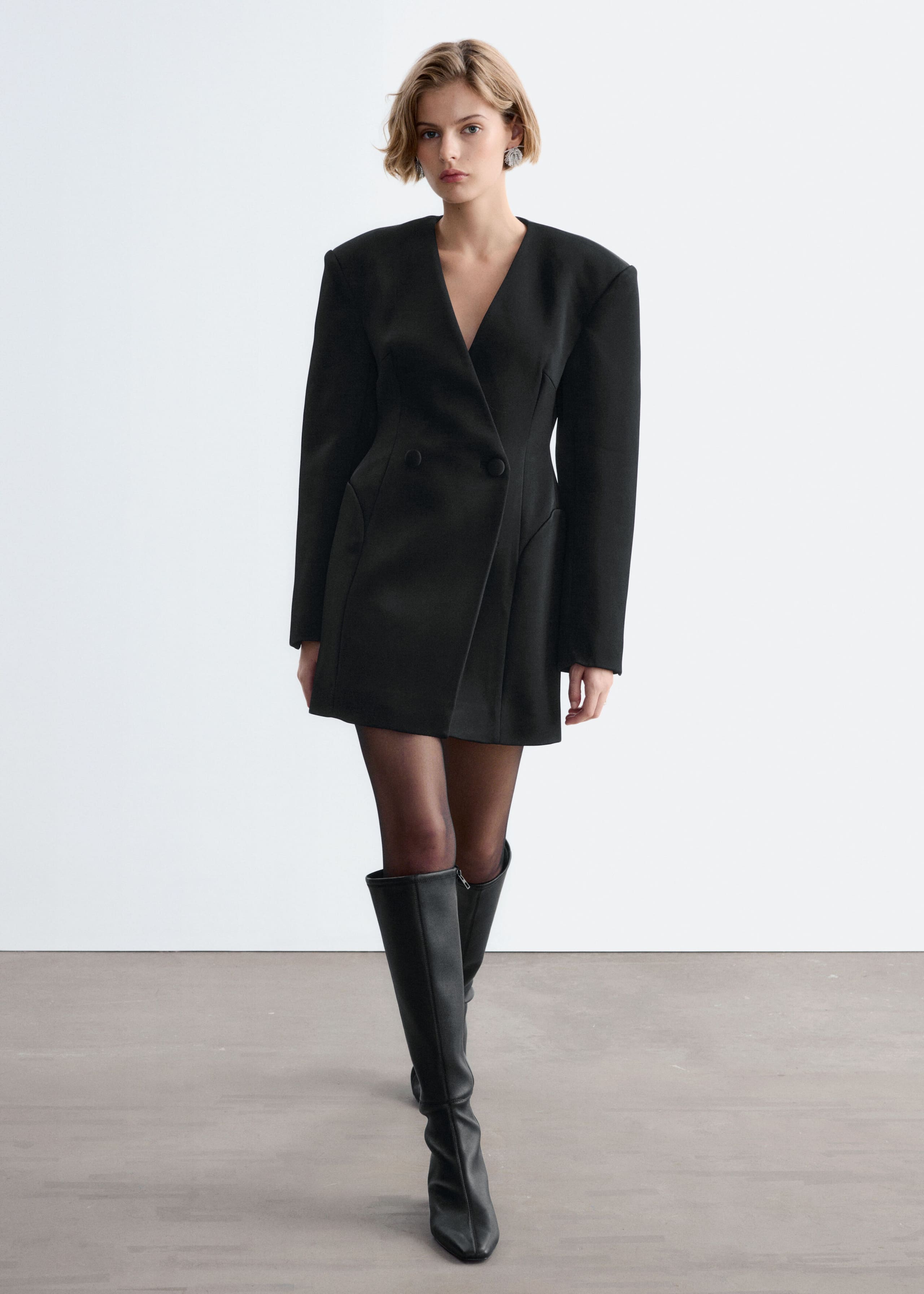 A black double-breasted blazer dress with structured shoulders, a deep V-neckline, and a cinched waist is worn by the model. The dress is styled with sheer black tights and knee-high black leather boots.