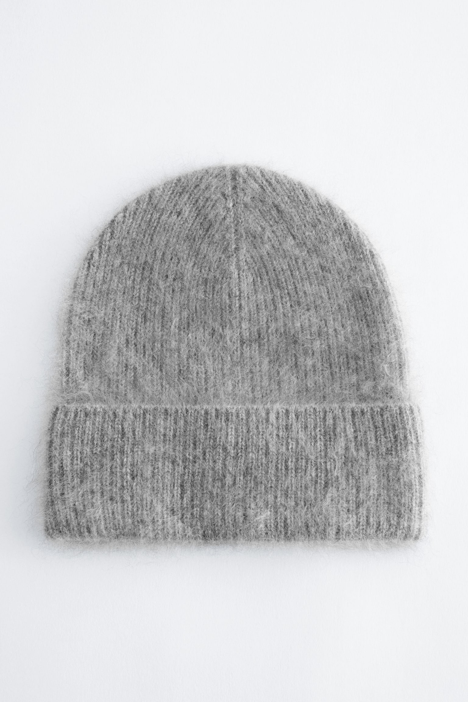 Un berretto lavorato a maglia grigio è mostrato frontalmente, con una finitura soffice e materica e una struttura a maglia a coste su tutta la superficie. Il cappello presenta un ampio risvolto ripiegato alla base.