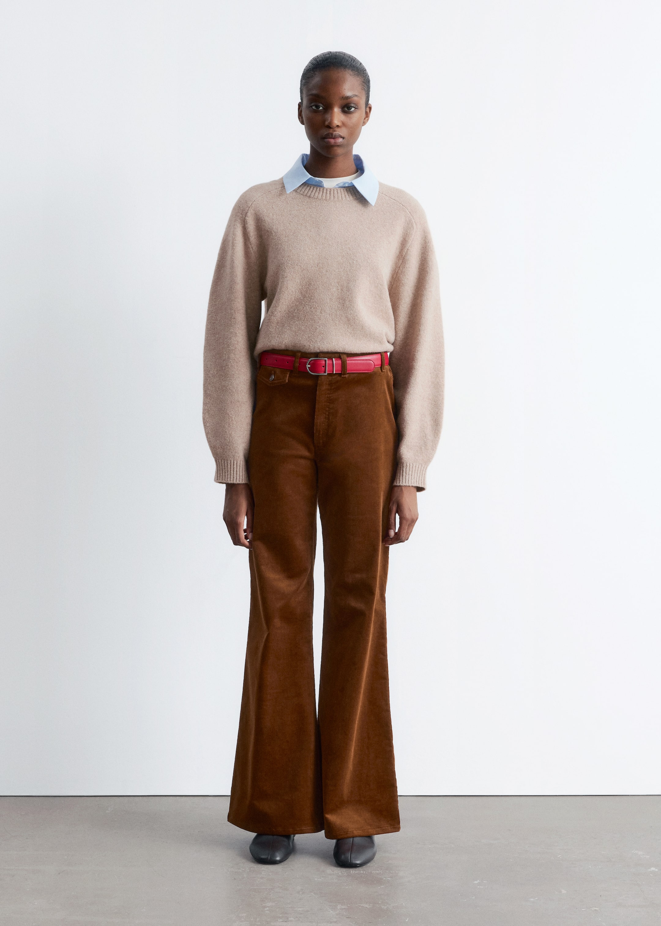 Grotere afbeelding bekijken: Een volledige outfit wordt frontaal gepresenteerd, met een beige trui met ronde hals en raglanmouwen over een lichtblauw overhemd met kraag. Een rode riem met een zilverkleurige gesp snoert bruine corduroy broek met wijde pijpen in. Donkere platte schoenen maken het ensemble compleet.