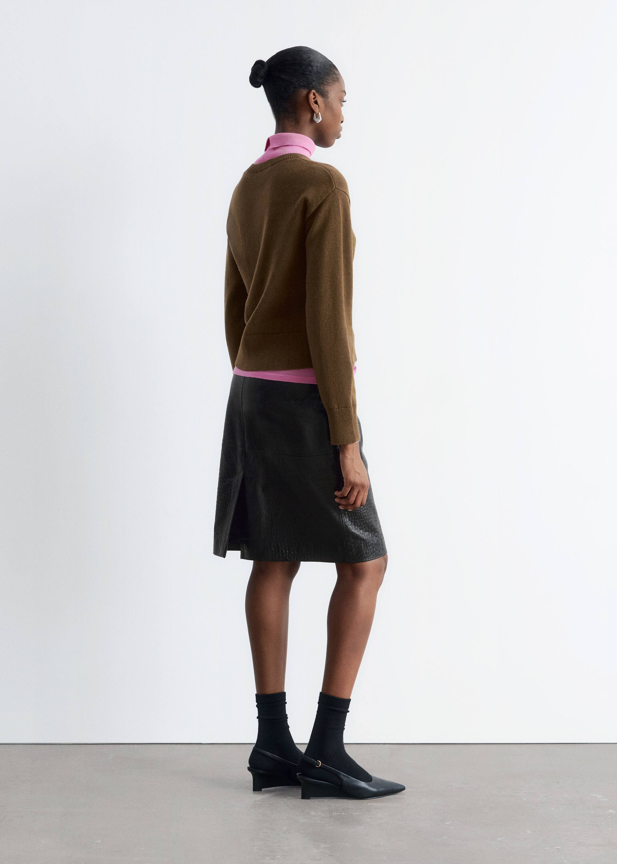 Se større billede: En person præsenterer et lagdelt outfit, vendt væk og let til højre. De har en brun langærmet sweater med ribbede manchetter og kant, lagdelt over en pink højhalset top. Outfittet er fuldendt med en sort knælængde nederdel med et subtilt tekstureret mønster og en slids bagpå, sorte ribbede crew-sokker og sorte slingback-sko med spids tå og en lav hæl.