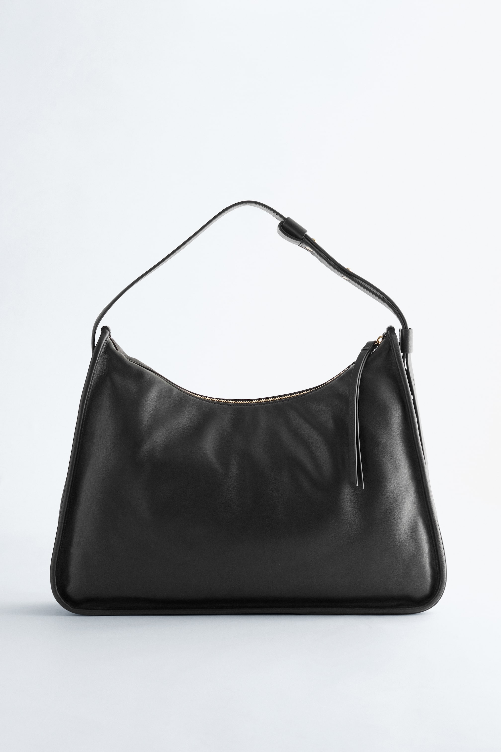 Ver imagen más grande: Un bolso de hombro negro con apariencia de cuero se presenta de FRENTE, con una forma suave y de media luna, y una única correa ajustable. Presenta un cierre de cremallera en tono dorado a lo largo de la parte superior curva, con dos finas lengüetas negras que se extienden desde el tirador de la cremallera.