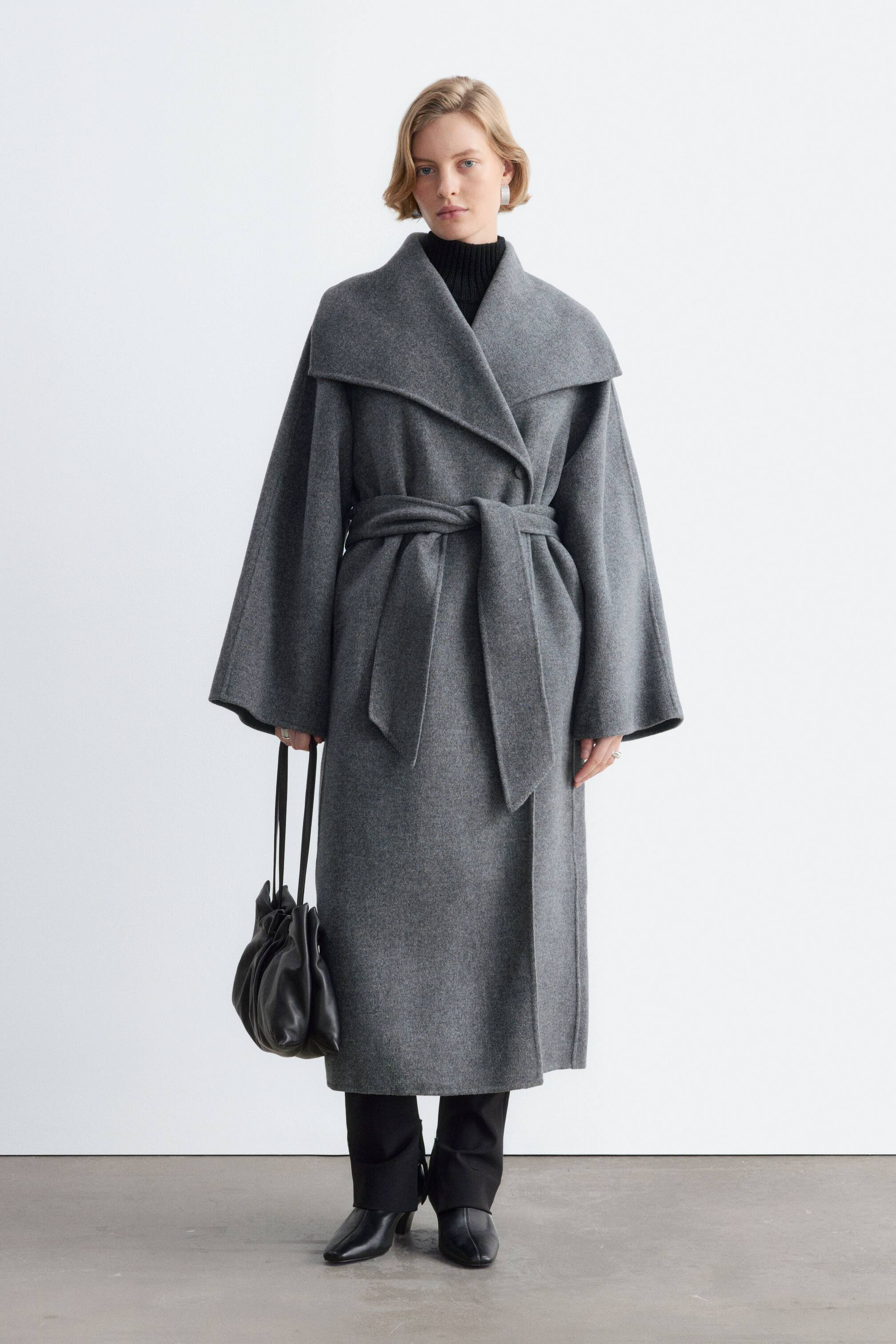 Visualizza l'immagine ingrandita: Un cappotto maxi in lana grigio, caratterizzato da un ampio colletto a scialle, maniche svasate e una cintura in vita abbinata da annodare, è indossato dalla modella. Il cappotto avvolgente è sovrapposto a un dolcevita nero e a pantaloni neri, mentre in una mano viene portata una borsa a mano in pelle nera.