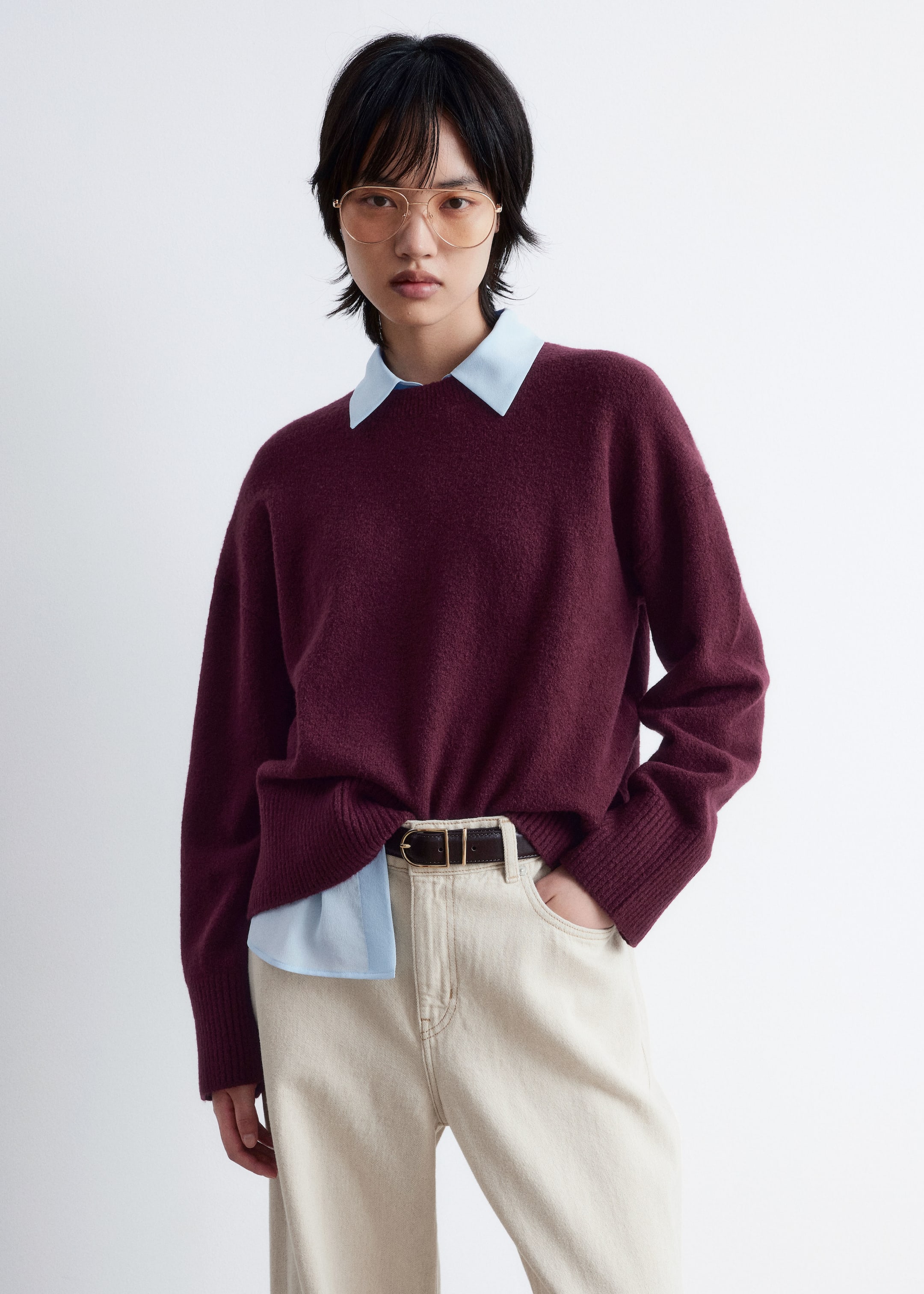 Grösseres Bild ansehen: Ein bordeauxroter Pullover mit Rundhalsausschnitt, der eine weiche Strickstruktur und eine lockere Passform aufweist, präsentiert lange Ärmel mit gerippten Bündchen. Sein gerippter Saum ist vorne teilweise eingesteckt und liegt über einem hellblauen Hemd mit Kragen, das am Halsausschnitt sichtbar ist.