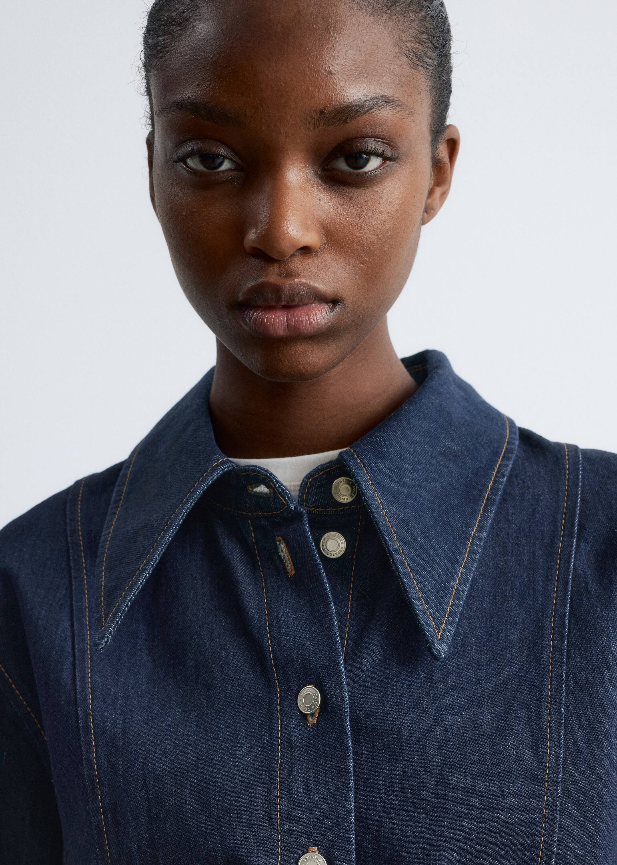 Ingrandisci l'immagine: Una camicia in denim indaco scuro è mostrata da un'angolazione frontale, con un ampio colletto a punta e bottoni a pressione argentati lungo l'abbottonatura anteriore. Cuciture a contrasto gialle dettagliano le cuciture e i bordi del colletto. Uno strato interno bianco è visibile sotto la camicia di denim.