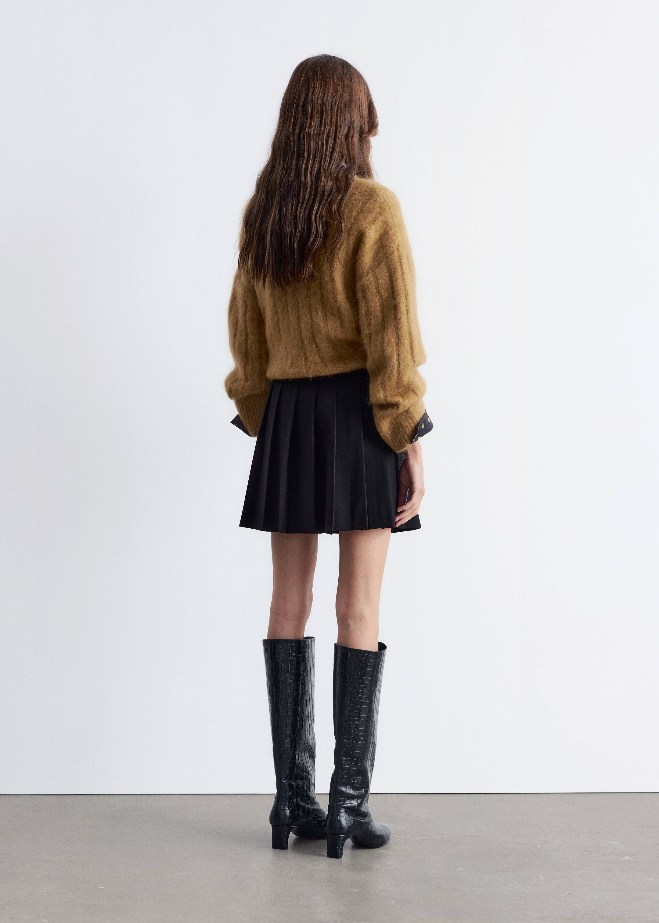 Ingrandisci l'immagine: Un maglione a coste di colore cammello, morbido e peloso, viene presentato dal retro, con maniche lunghe risvoltate per rivelare i polsini a pois di un indumento scuro sottostante. È abbinato a una mini-gonna nera a pieghe e stivali neri al ginocchio con una texture in rilievo e tacchi a blocco.