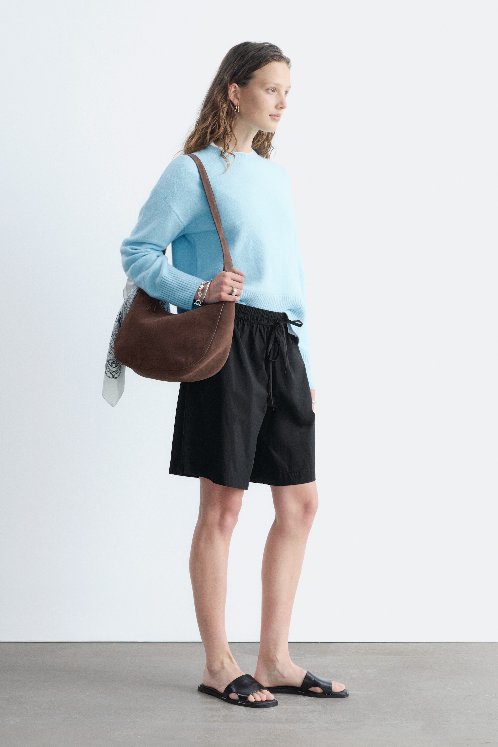 Grösseres Bild ansehen: Ein hellblauer Pullover mit Rundhalsausschnitt präsentiert sich in einer entspannten Passform mit langen Ärmeln, gerippten Bündchen und einem gerippten Saum, aus einem seitlich-vorderen Winkel betrachtet. Eine braune Wildleder-Hobo-Tasche ruht über der Vorderseite und ein gemustertes Tuch ist an der Hüfte sichtbar.
