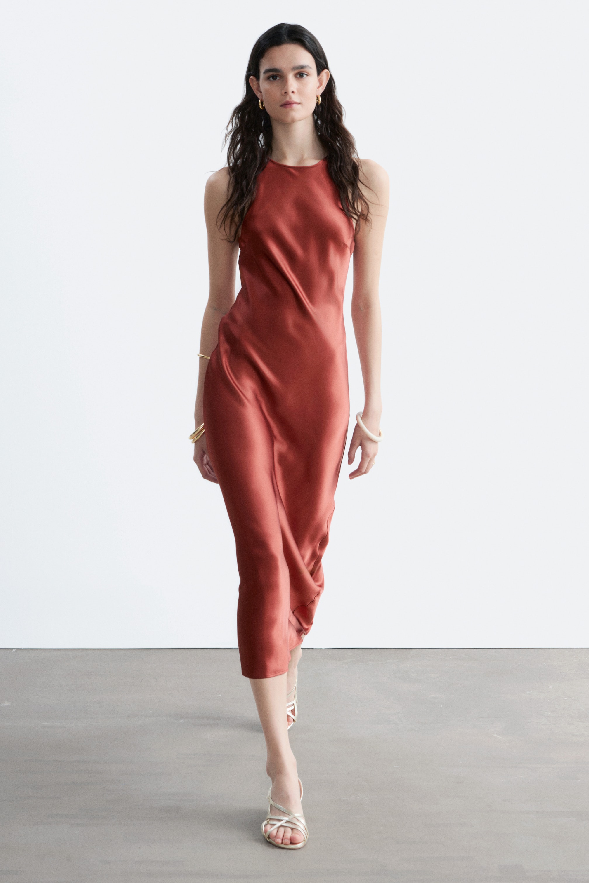 Ver imagen más grande: Un vestido midi de tirantes color óxido en tela de acabado satinado, que presenta un escote alto y una silueta fluida que se ajusta al cuerpo, mientras se muestra desde un ángulo ligero.