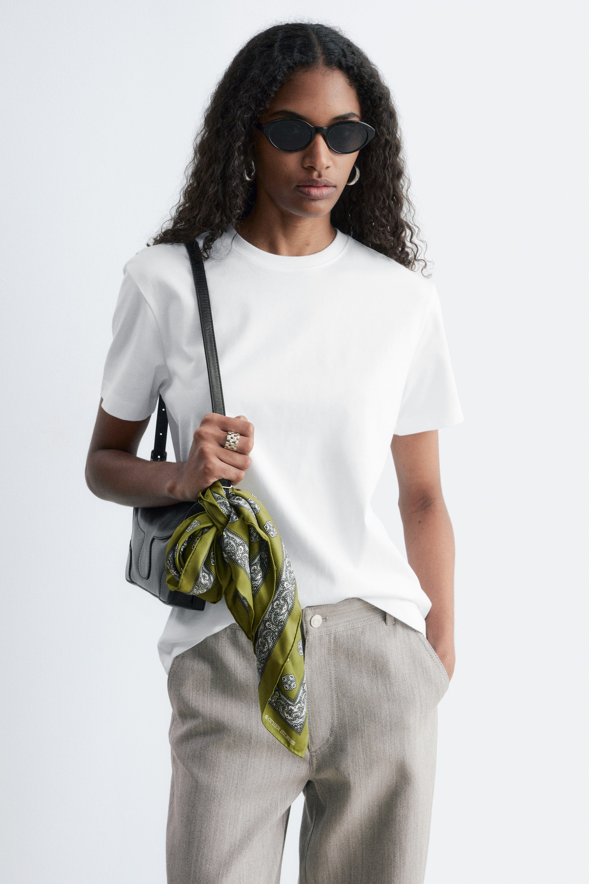 Größeres Bild anzeigen: Ein weißes T-Shirt mit kurzen Ärmeln, das einen Rundhalsausschnitt und einen glatten Stoff aufweist, präsentiert sich in einer entspannten Passform. Es wird getragen, wobei eine Seite in hellfarbige Hosen gesteckt ist, und ein schwarzer Schulterriemen einer Tasche kreuzt sich über die Vorderseite.