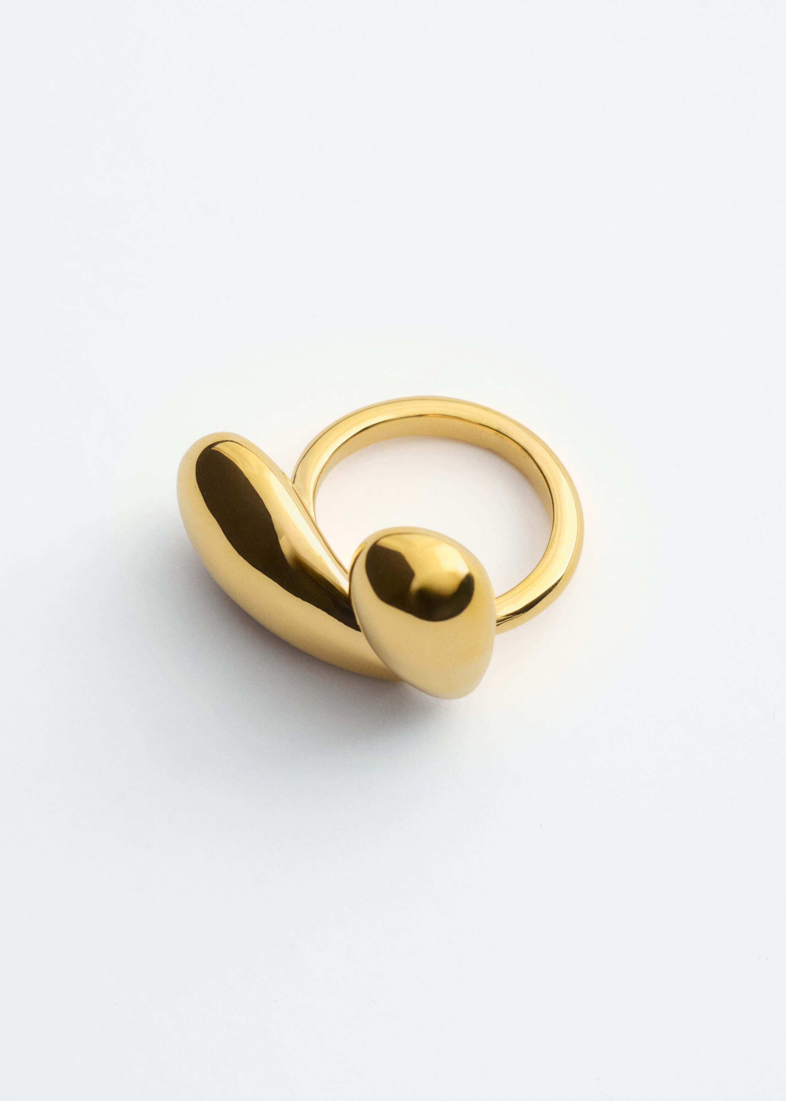 Un anello in ottone lucido presenta un design scultoreo definito da due forme fluide e arrotondate con una finitura altamente riflettente. L’anello è ripreso frontalmente da un’angolazione rialzata, mettendo in evidenza come gli elementi lisci a forma di goccia si incurvino sopra la fascia circolare.
