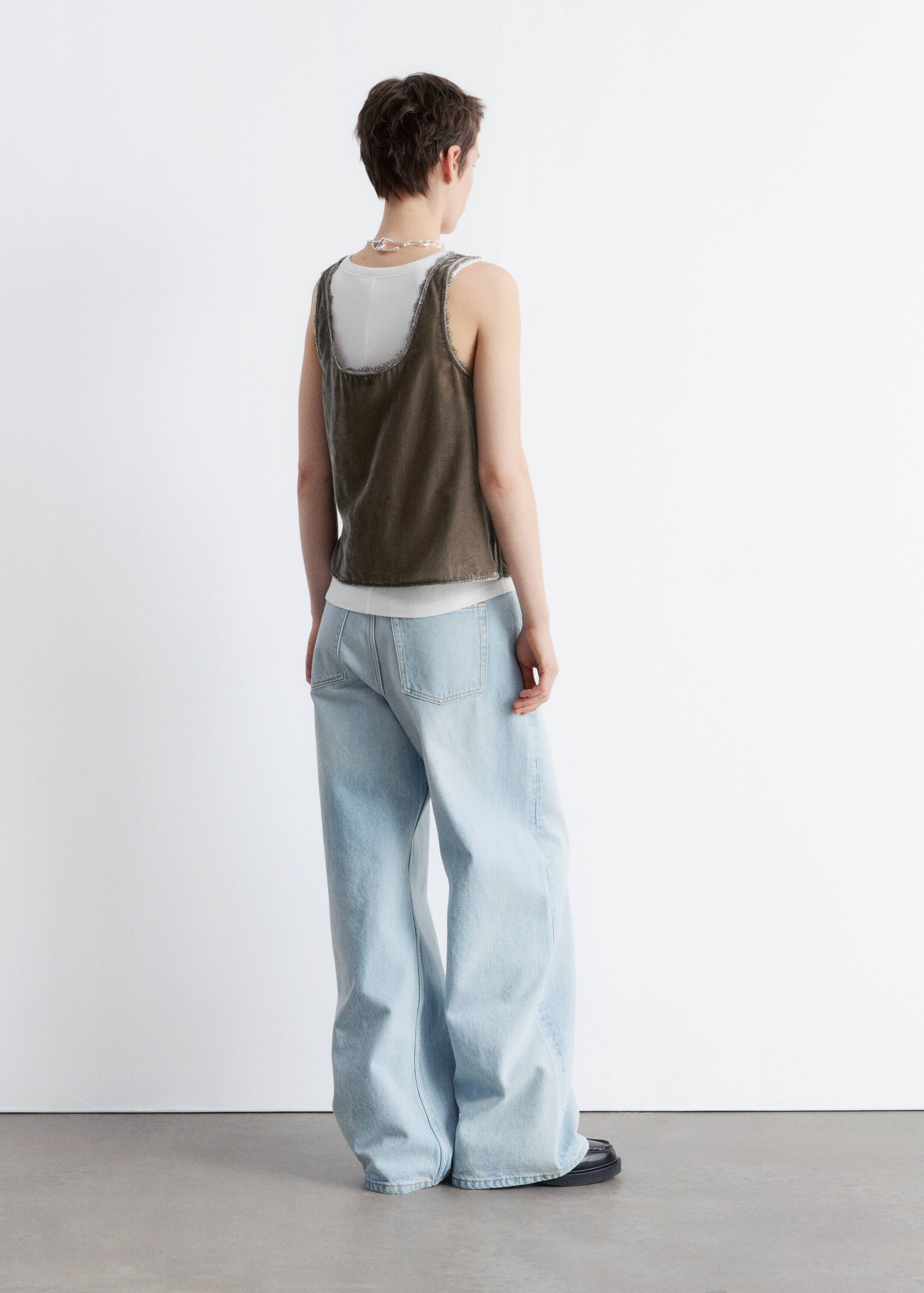 Größeres Bild anzeigen: Ein dunkles, ärmelloses, kurzes Oberteil mit abgenutzten Kanten wird von hinten gezeigt, über einem weißen gerippten Tanktop. Es wird mit hellblauen, weit geschnittenen Jeans kombiniert, die an den Knöcheln zusammenfallen.