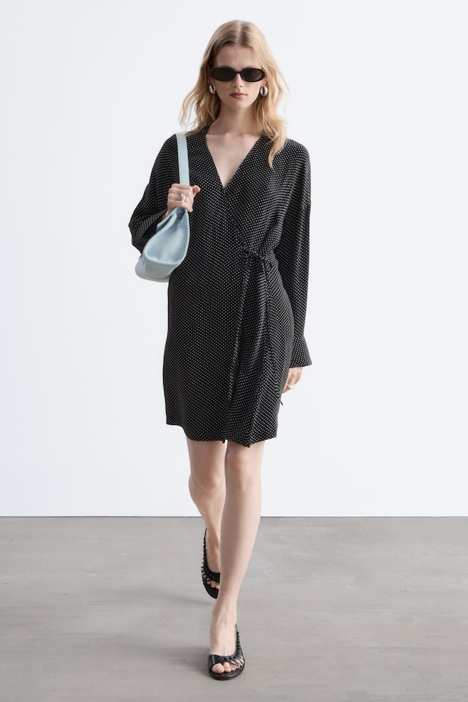 Een zwarte wikkeljurk met stippen heeft een V-hals en lange mouwen, met een zelfbindende sluiting die de taille accentueert. De jurk is gestyled met een lichtblauwe schoudertas en zwarte open schoenen. - 1