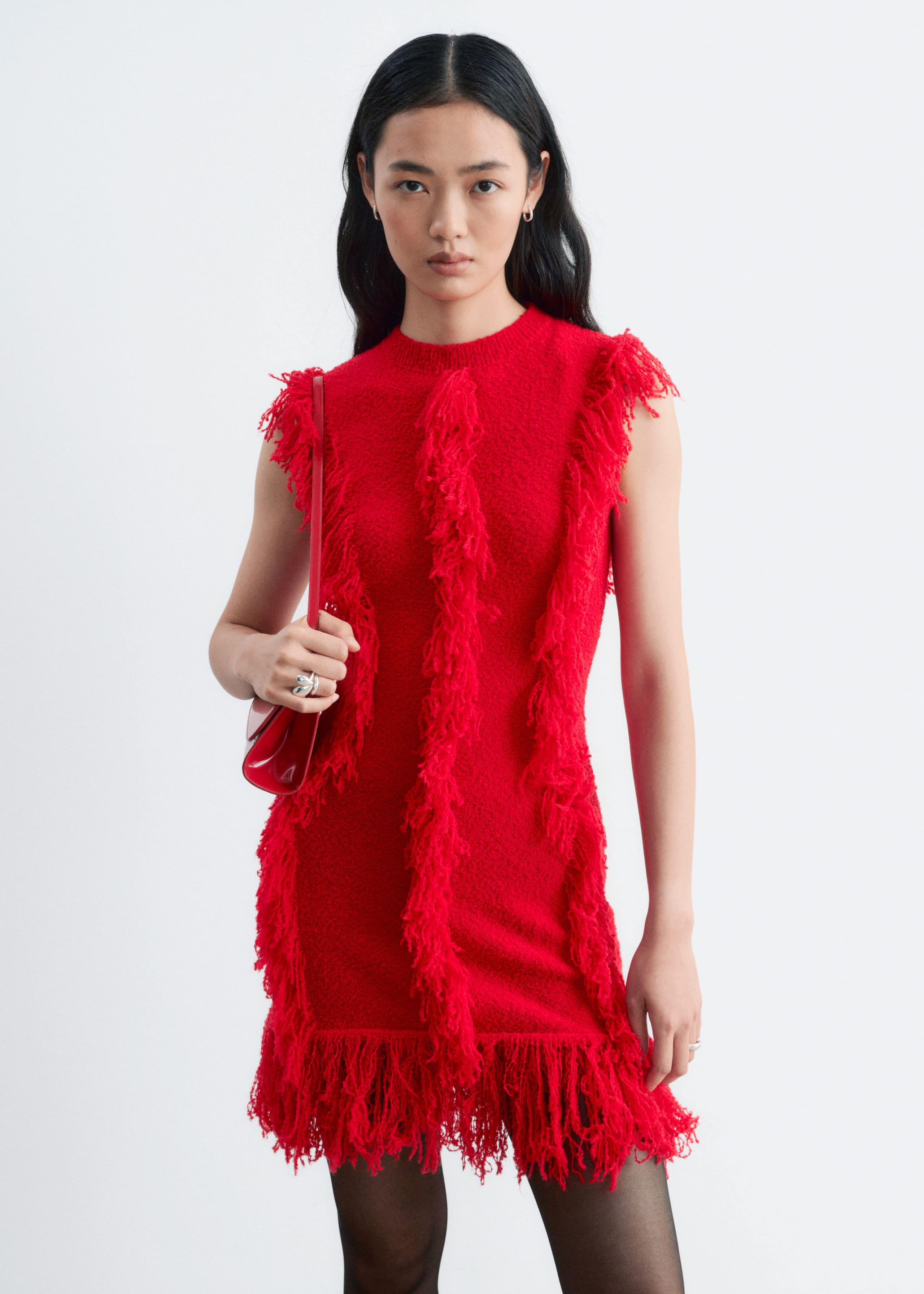 A red, sleeveless knitted dress featuring vertical rows of textured fringe and a heavily fringed hem is being worn by the model. The model is styling the dress with black sheer tights and carrying a matching red shoulder bag.