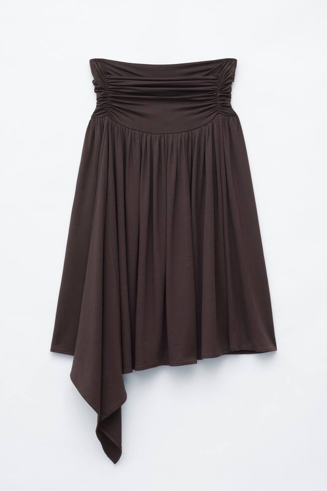 Uma saia midi em jersey franzido castanho-escuro Ã© apresentada de frente, apresentando uma cintura alta com franzido horizontal que transita para uma estrutura drapeada e fluida. A saia cai em cascata com uma bainha assimÃ©trica, realÃ§ada por um painel de tecido mais comprido e pendente no lado inferior esquerdo. - 1