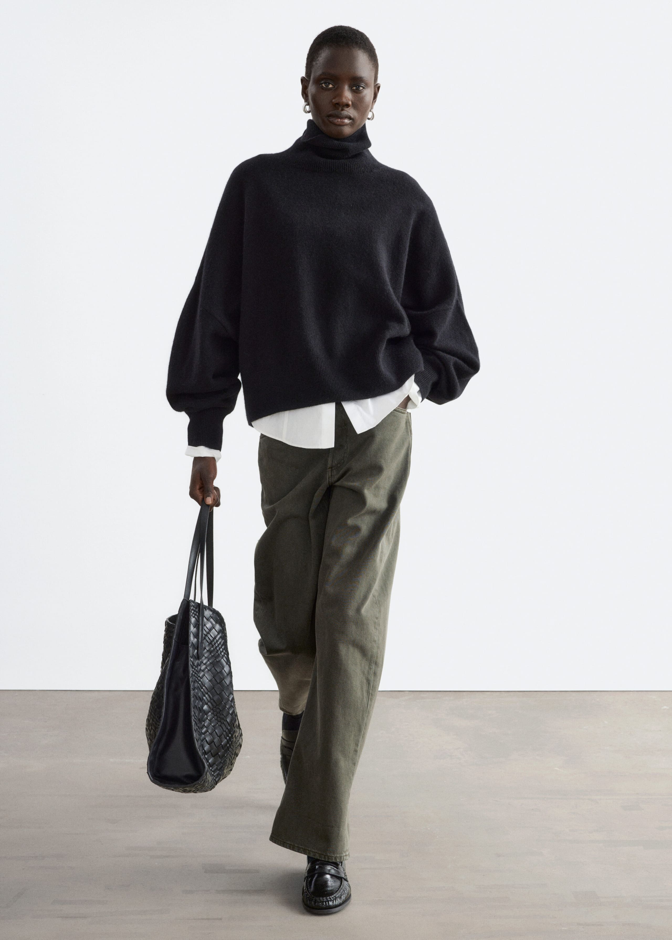 A black, oversized knitted roll-neck jumper featuring dropped shoulders and ribbed cuffs is worn by the model. The jumper is layered over a white collared shirt and paired with olive-green wide-leg trousers.