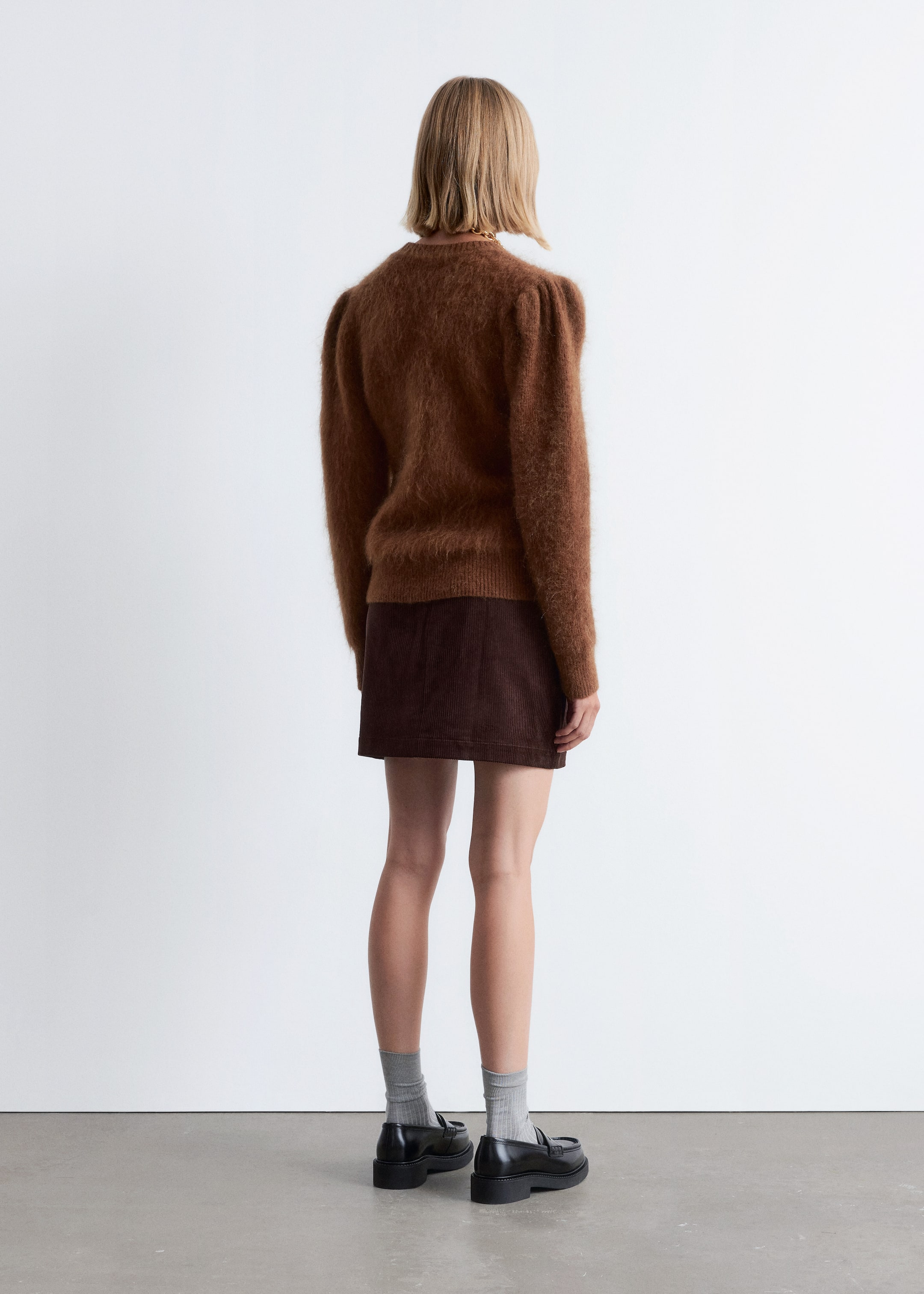 Visualizza immagine più grande: Un maglione lavorato a maglia marrone sfocato con spalle a sbuffo e orlo a coste viene mostrato da dietro. Una minigonna in velluto a coste marrone scuro, con costine verticali visibili e un taglio dritto, viene indossata sotto.