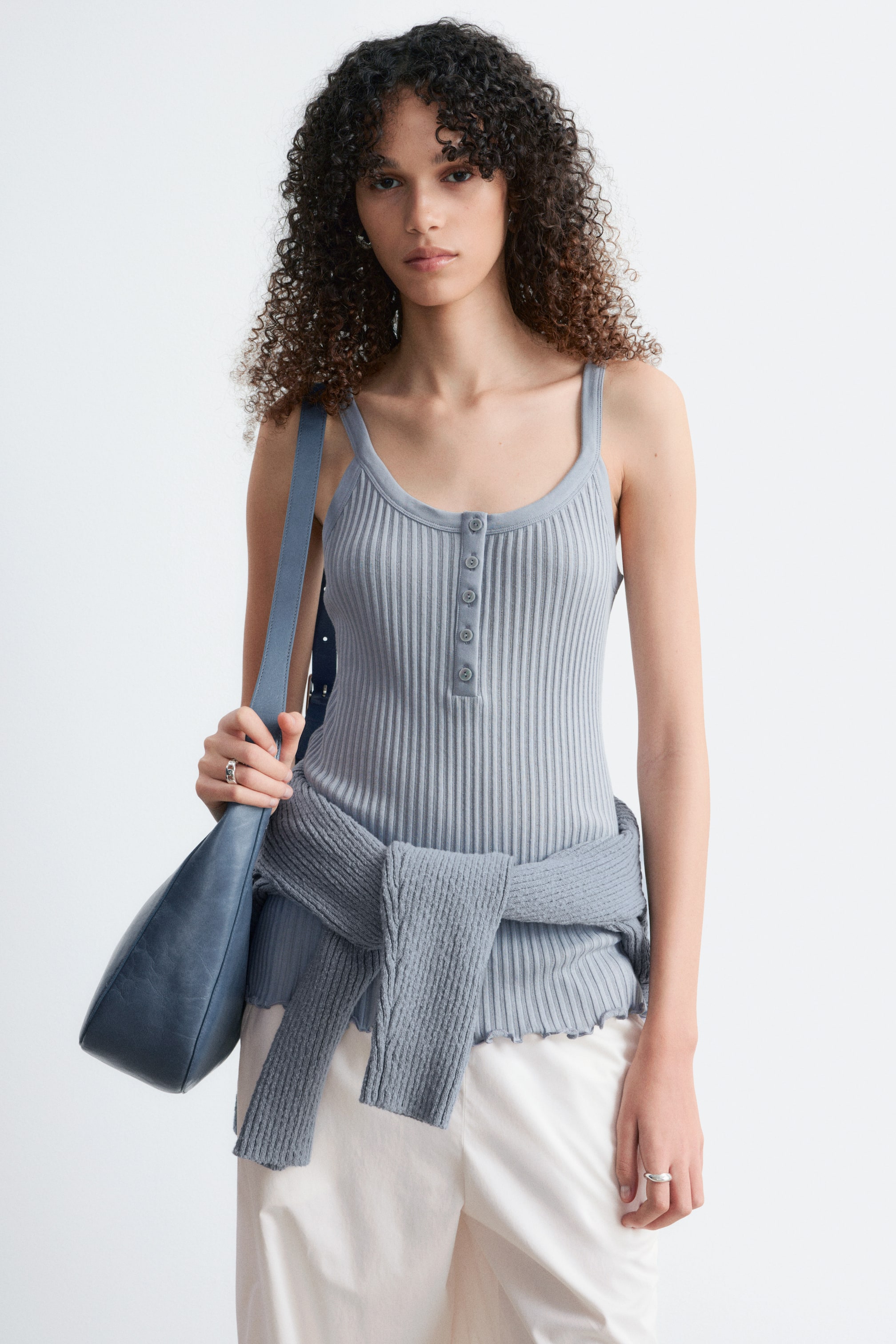 Zobrazit větší obrázek: Světle modré žebrované tílko s tenkými ramínky, kulatým výstřihem a středovou knoflíkovou légou. Kolem pasu je uvázaný odpovídající žebrovaný pletený svetr, který doplňuje vlnitý okraj tílka ve stylu "salátového listu".