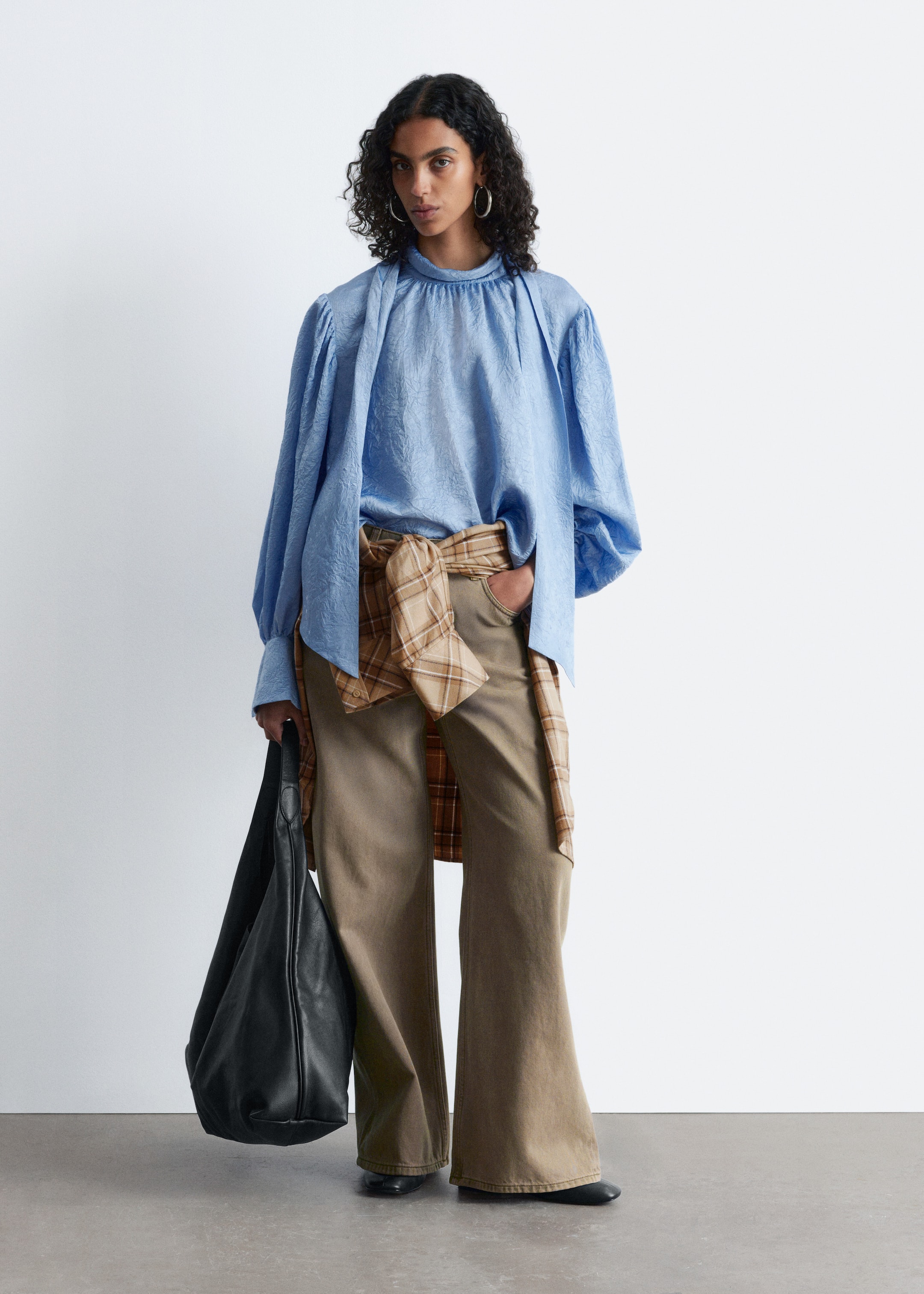 Visualizza immagine più grande: Una camicetta azzurra testurizzata, con un alto scollo arricciato e maniche voluminose con polsini aderenti, è indossata con pantaloni ampi verde oliva. Una camicia a quadri è annodata in vita e una borsa nera morbida è portata a mano, completando l'ensemble frontale.