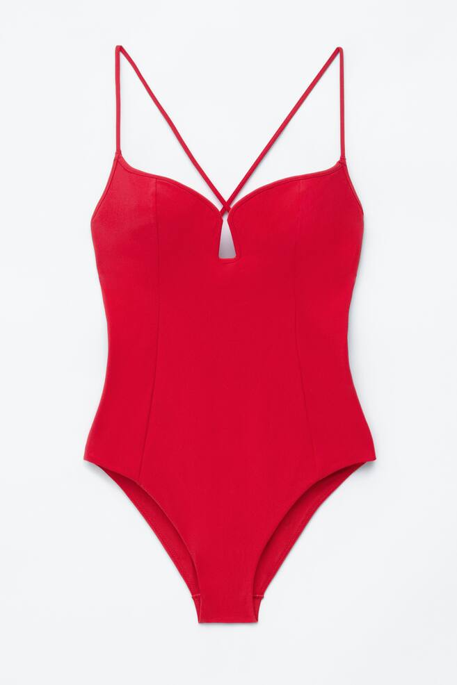 A red swimsuit is lying flat, viewed from the front. It features a triangle-cut neckline with a small central cut-out, and slim straps originate from the shaped cups, crossing over the chest. Vertical seams define the torso. - 1