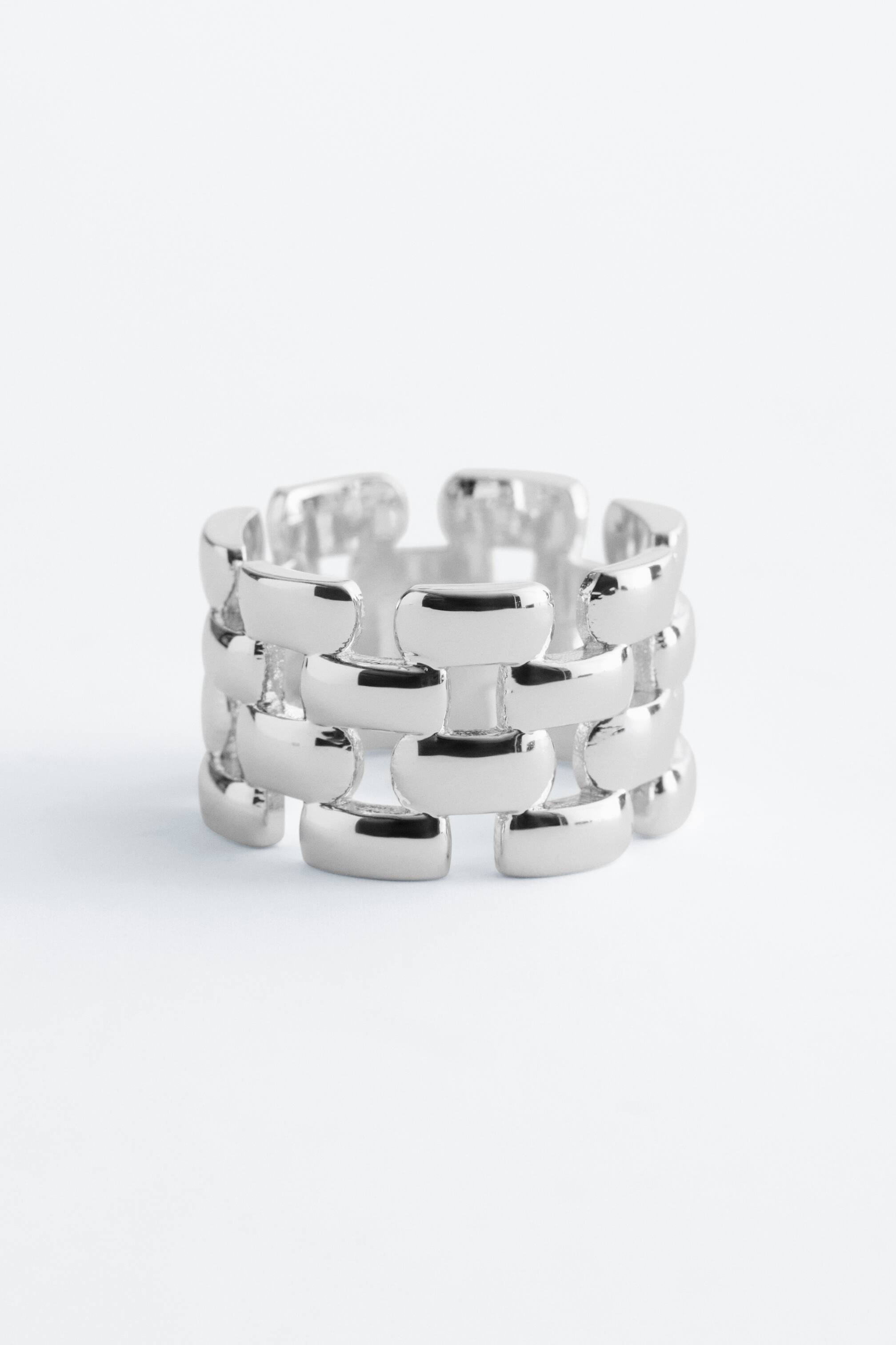 Grotere afbeelding bekijken: Een stevige, zilverkleurige ring, vervaardigd uit gepolijst messing, toont een architecturaal kettingpatroon van in elkaar grijpende vierkante schakels. Het reflecteert licht en is gepositioneerd in een iets schuin vooraanzicht.