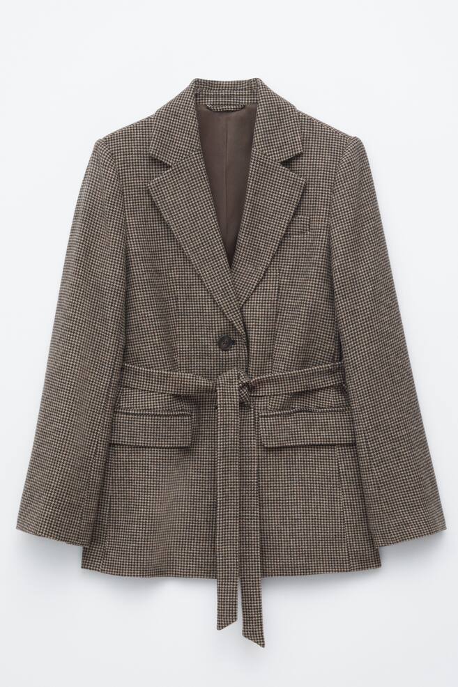 A single-breasted blazer featuring a brown houndstooth pattern is hanging facing front, showcasing notch lapels and a single-button fastening. The blazer is designed with a matching tie belt cinched at the waist, long sleeves, and two front flap pockets. - 1