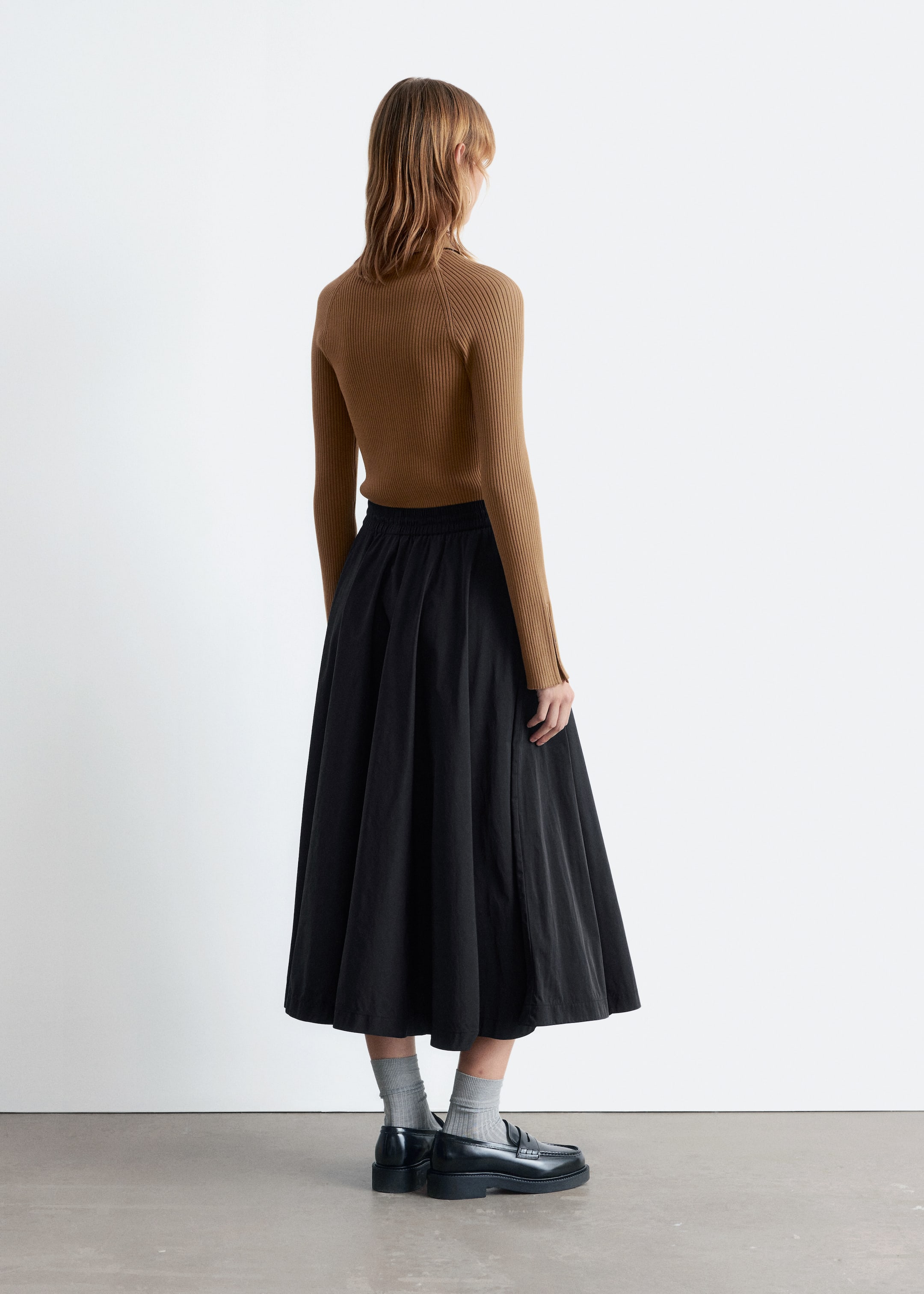 Agrandir l'image: Une jupe midi noire est présentée sous un angle arrière-droit, avec une silhouette ample et évasée et une taille élastiquée. Le tissu de la jupe tombe en plis souples juste au-dessus des chevilles, et elle est portée avec un haut marron côtelé ajusté rentré, mettant en valeur la taille froncée de la jupe.