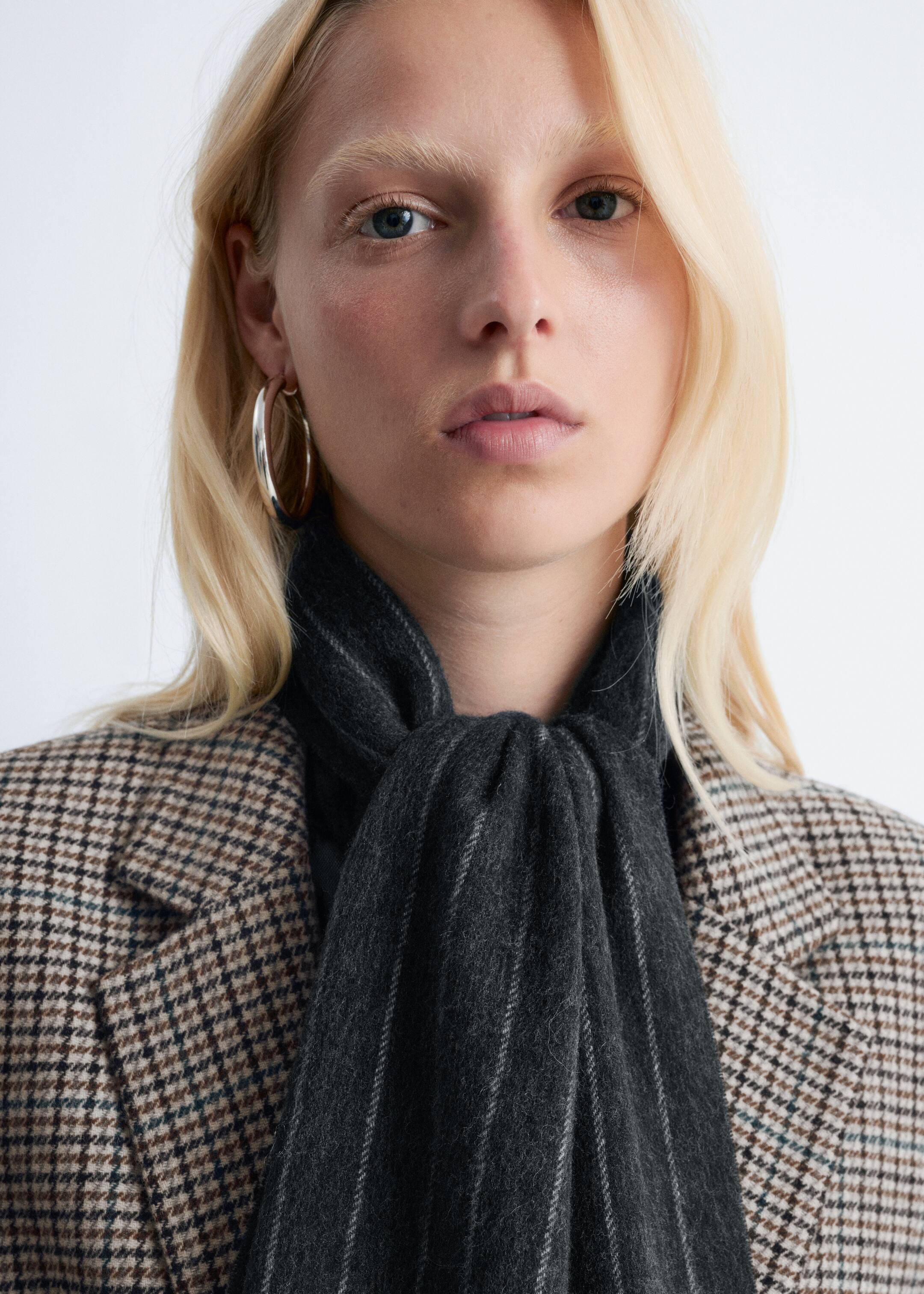 Größeres Bild anzeigen: Ein dunkelgrauer Nadelstreifen-Schal wird um den Hals des Models gewickelt, wobei seine Enden hinunterhängen. Der Schal ist über einem braun-schwarzen karierten Blazer gestylt.